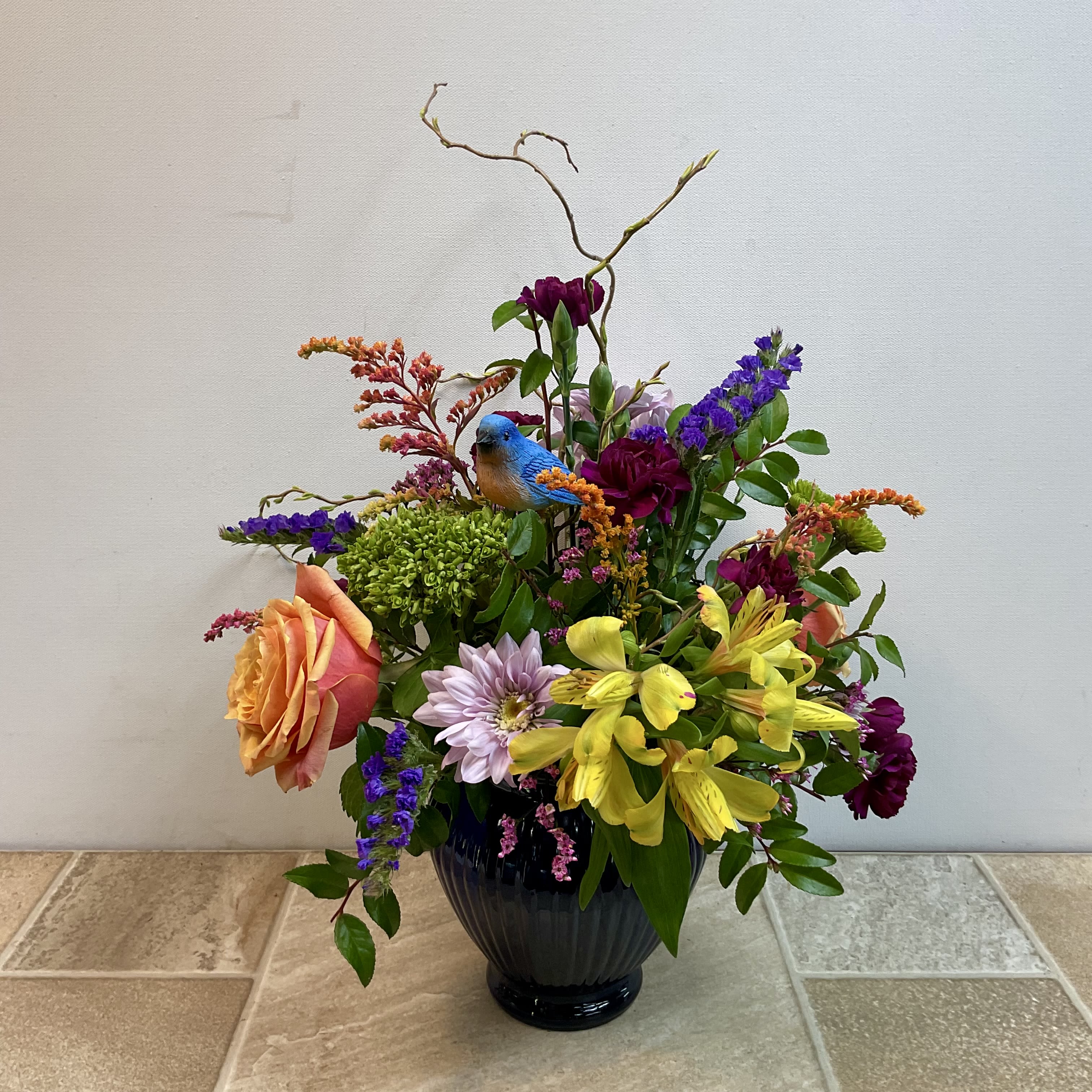 Songbird Serenade - A beautiful arrangement filled with a bright and cheerful collection of our favorite fresh flowers. It includes lavender daisies, yellow alstroemeria, cherry brandy roses and much more arranged in a pretty blue glass vase. It features a lovely ceramic bluebird nestled within the blooms and measures 12” wide x 14” tall.
