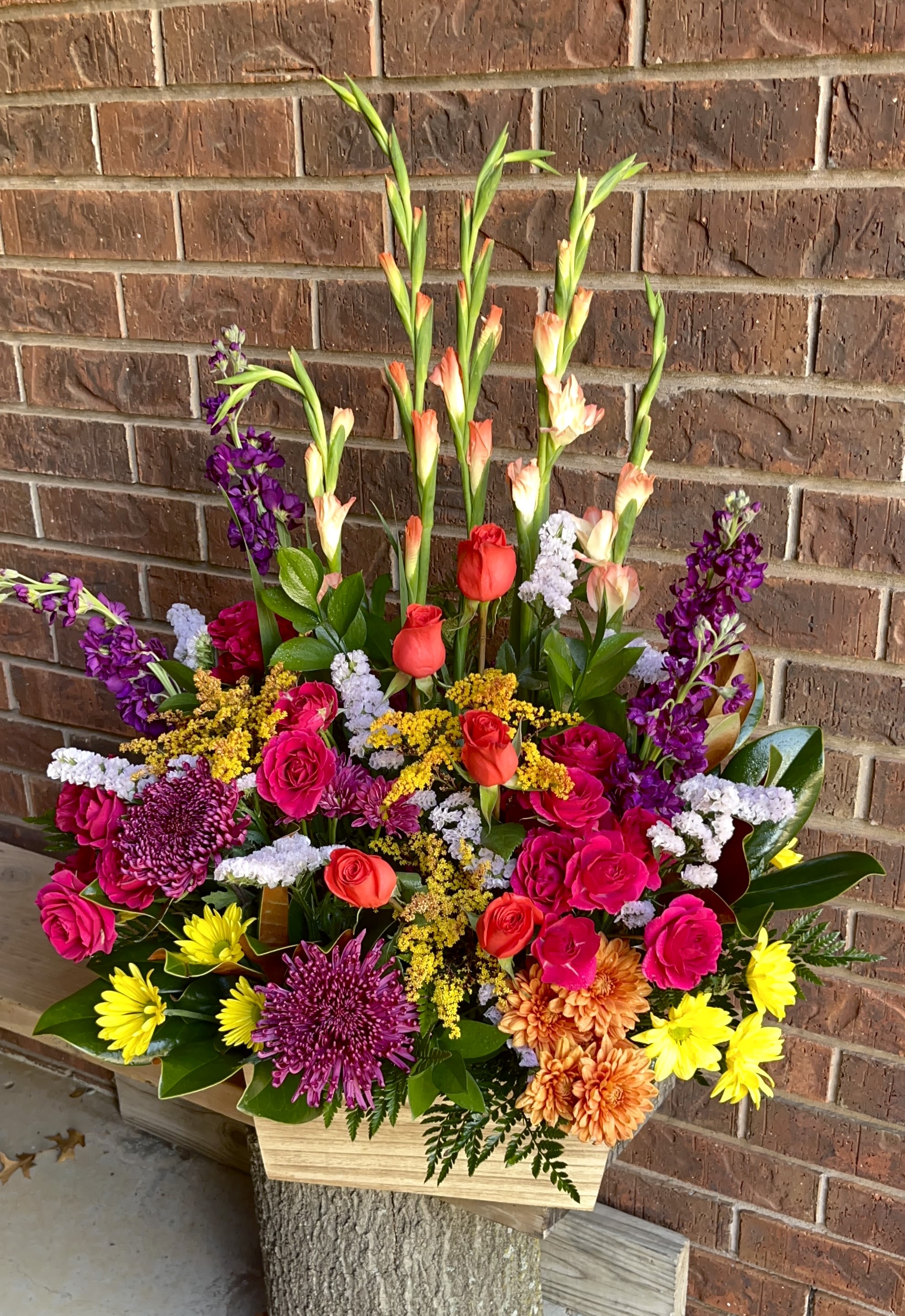 Sunset Tribute Basket - Bright pink roses and red spray roses mingle with purple chrysanthemums and sunny yellow daisy-like blooms in this Sunset Tribute Basket, a vibrant flower arrangement perfect for sympathy, congratulations, or any celebratory moment. The garden-style mix reads joyful and generous-designed to make the person you're thinking of feel truly celebrated with a warm, colorful impact that catches the eye from across the room.  Arranged densely in a low wooden container, the bouquet features layered textures: velvety roses, ruffled carnations and chrysanthemums, classic daisies, and taller peach-and-green spike flowers that give uplifting vertical accents above the main body. The abundant, multicolor palette-pink, purple, yellow and red-creates a lively contrast while the wooden vessel adds rustic charm and stability. The overall size reads large and full, crafted to command attention on a table or mantel.  Ideal for brightening a special day, honoring a loved one,  or saying "well done," this arrangement suits recipients who love cheerful, garden-inspired florals. No-fuss care: keep flowers in fresh water, out of direct sun, and trim stems if needed to maintain freshness; available in the pictured large design for an impressive presentation.