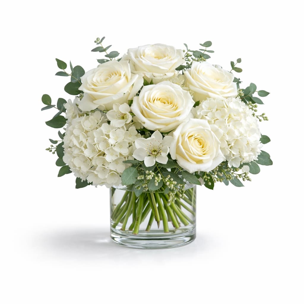 Cloud Nine Blooms - Creamy white roses nestled above rounded white hydrangea blooms give this flower arrangement a serene, refined presence-perfect as a sympathy arrangement, anniversary roses, or a calming birthday bouquet. The soft monochromatic palette of white and green reads as quietly romantic and will make the recipient feel gently seen and comforted.  Compact and full in shape, the design pairs dense hydrangea heads with clustered roses, airy eucalyptus-like foliage, and delicate white filler flowers for texture and movement. Arranged in a clear glass vase, the composition keeps a clean, classic silhouette that suits both everyday gifting and formal settings.  Choose this calming bouquet for someone who appreciates understated elegance-a thoughtful way to say you're thinking of them, congratulate a milestone, or offer condolences. The arrangement arrives ready to display in its vase; to keep blooms fresh, change the water every few days and trim stems at an angle when refreshing the display.