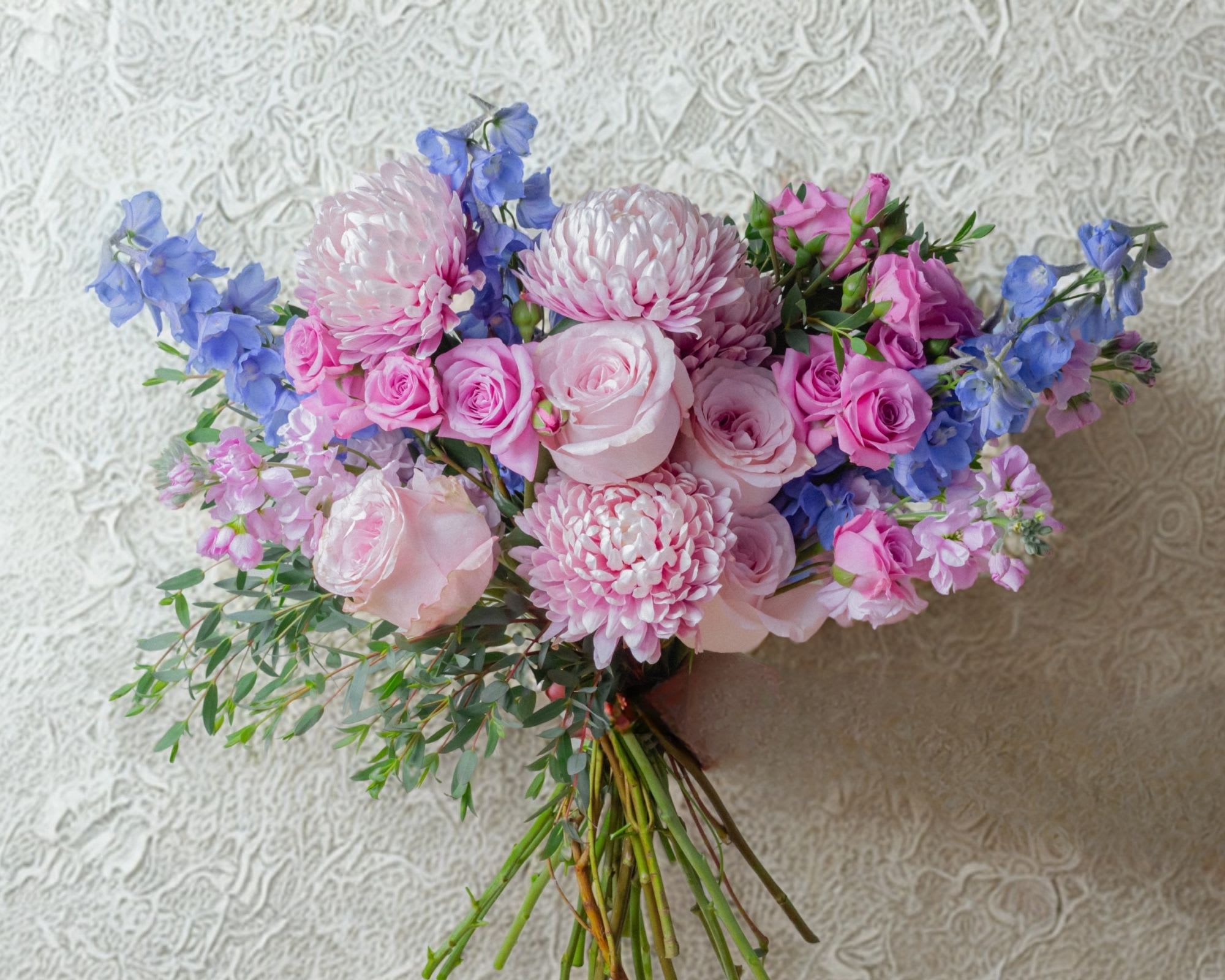 Blue Lavender Floral Bouquet - A dreamy blend of blush and pastel tones, this hand-tied bouquet features lush pink roses, full chrysanthemum blooms, and delicate accents of soft blue delphinium. Light, airy greenery adds movement and a natural garden feel, creating a perfectly balanced arrangement that feels both fresh and timeless.  Designed with a loose, organic style, this bouquet is ideal for birthdays, anniversaries, baby showers, or simply brightening someone’s day. Its gentle color palette and textural variety make it a standout piece that feels luxurious yet approachable.  Vase available to be added. 