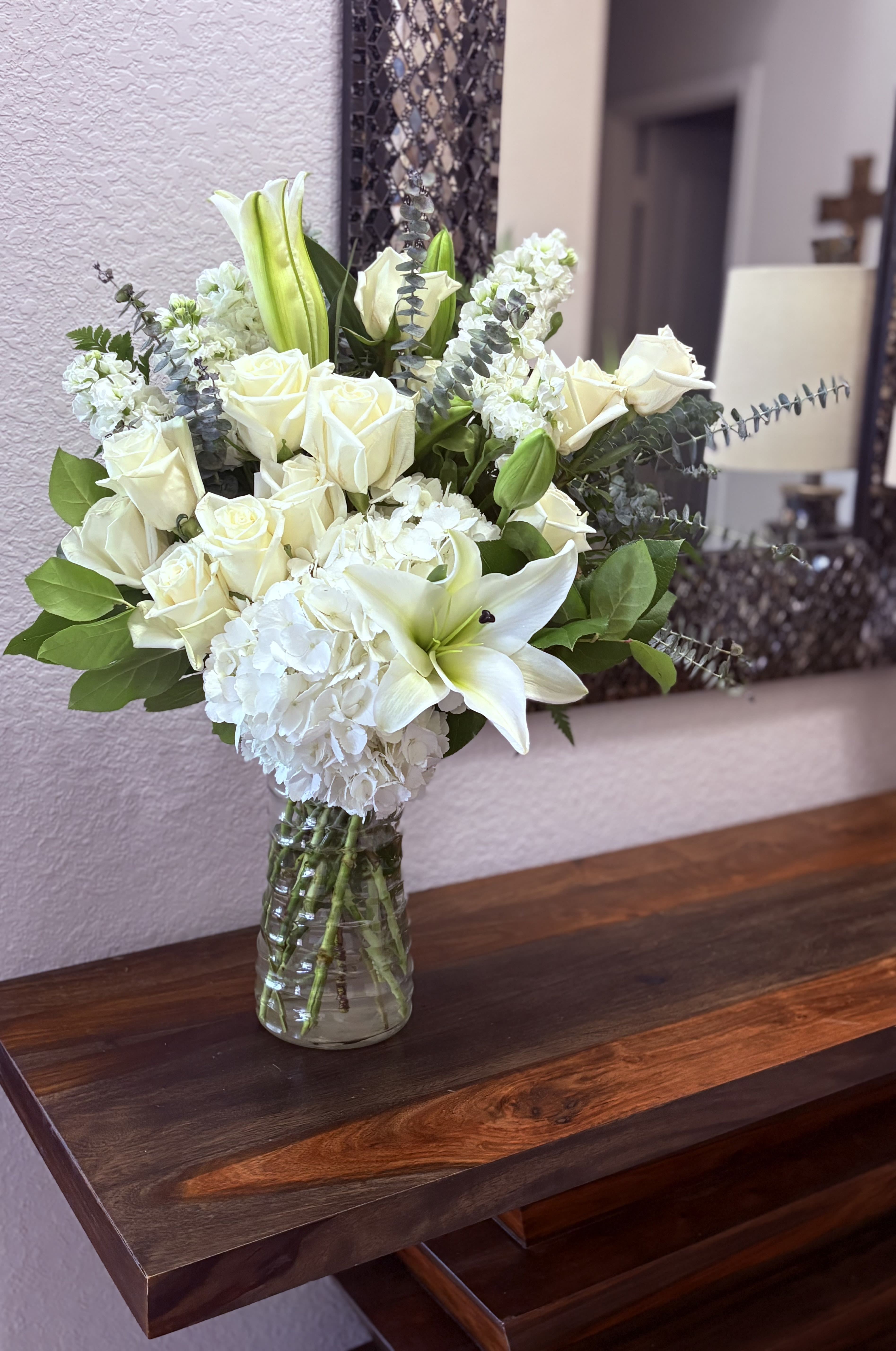 Love luxe Bouquet - Creamy white roses and a single white lily take center stage in this Love luxe bouquet, a refined flower arrangement perfect for a wedding, sympathy tribute, or an elegant everyday gift. The mostly white palette with muted green accents creates a calm, formal impression that will make the person you're thinking of feel comforted and truly seen.  A large cluster of white hydrangea gives the design its lush, rounded silhouette while soft green buds and silvery eucalyptus add vertical texture and gentle movement. Arranged in a clear glass vase, the bouquet reads fresh and sophisticated-clean lines, full volume, and a serene color story that complements both traditional and modern settings.  Choose this arrangement when you want a quietly luxurious gesture-whether to honor a milestone, express sympathy, or elevate a special table. The compact, balanced shape fits well on dining or console tables; keep flowers in fresh water, trim stems occasionally, and display out of direct heat for longest enjoyment.