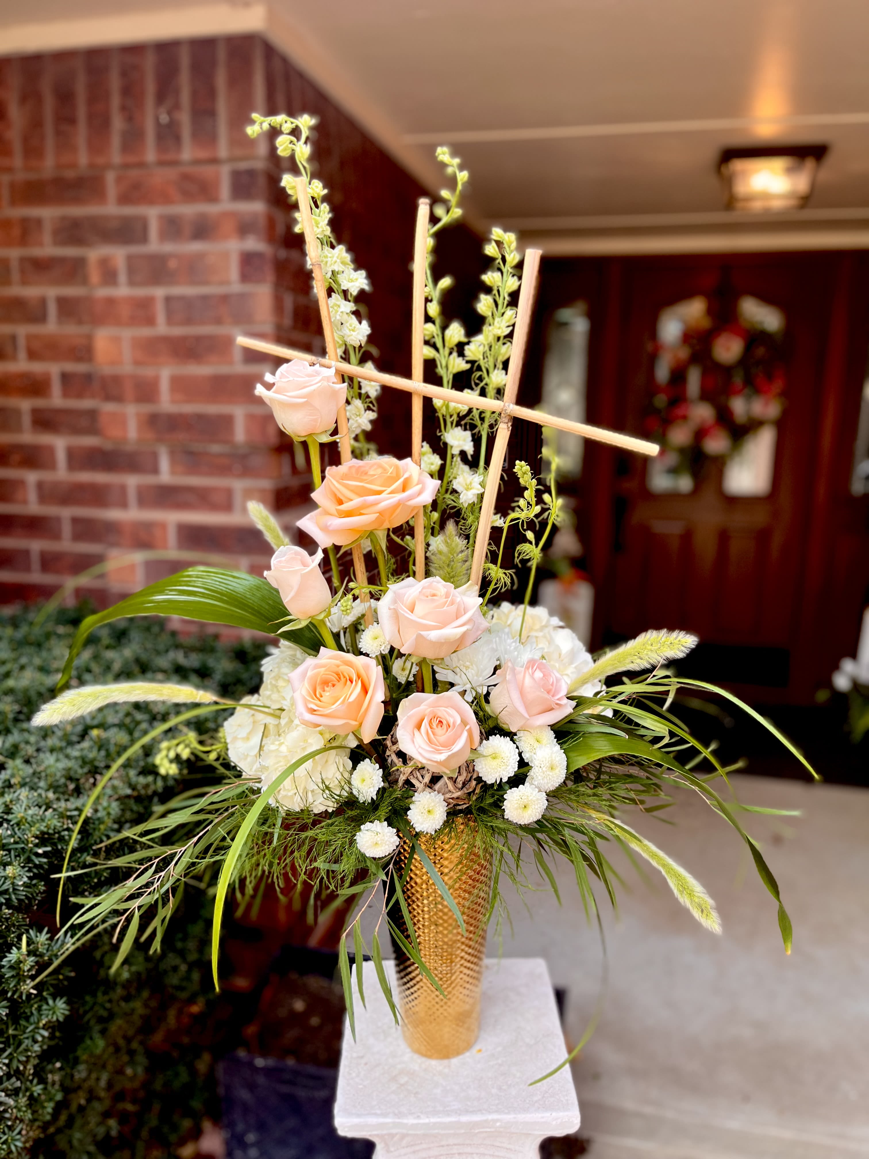 Peach Trellis - Peach roses gathered into a vertical, sculptural display make an immediate statement - a peach bouquet perfect for a wedding, anniversary, or any celebratory event where elegance matters. The Peach Trellis arrangement uses the warm glow of peach roses and soft white blooms to create a refined focal piece that will make the person you're honoring feel truly celebrated.  This tall arrangement is built on a gold-textured vase with bamboo-like wooden stakes and a white pedestal stand for added height and formality. Peach roses are clustered amid airy green stems, long grass blades, and small white chrysanthemums and filler flowers, with hints of yellow tones that brighten the composition. The design reads modern and structured: clean vertical lines, contrasting textures, and a subtle, sophisticated color palette.  Ideal as a ceremony centerpiece, entryway statement, or gift to mark a milestone, Peach Trellis sends a clear message of thoughtful celebration. Arrange on a pedestal or table where the height can be appreciated; water upon delivery and keep away from direct heat to help the roses and chrysanthemums last longer.
