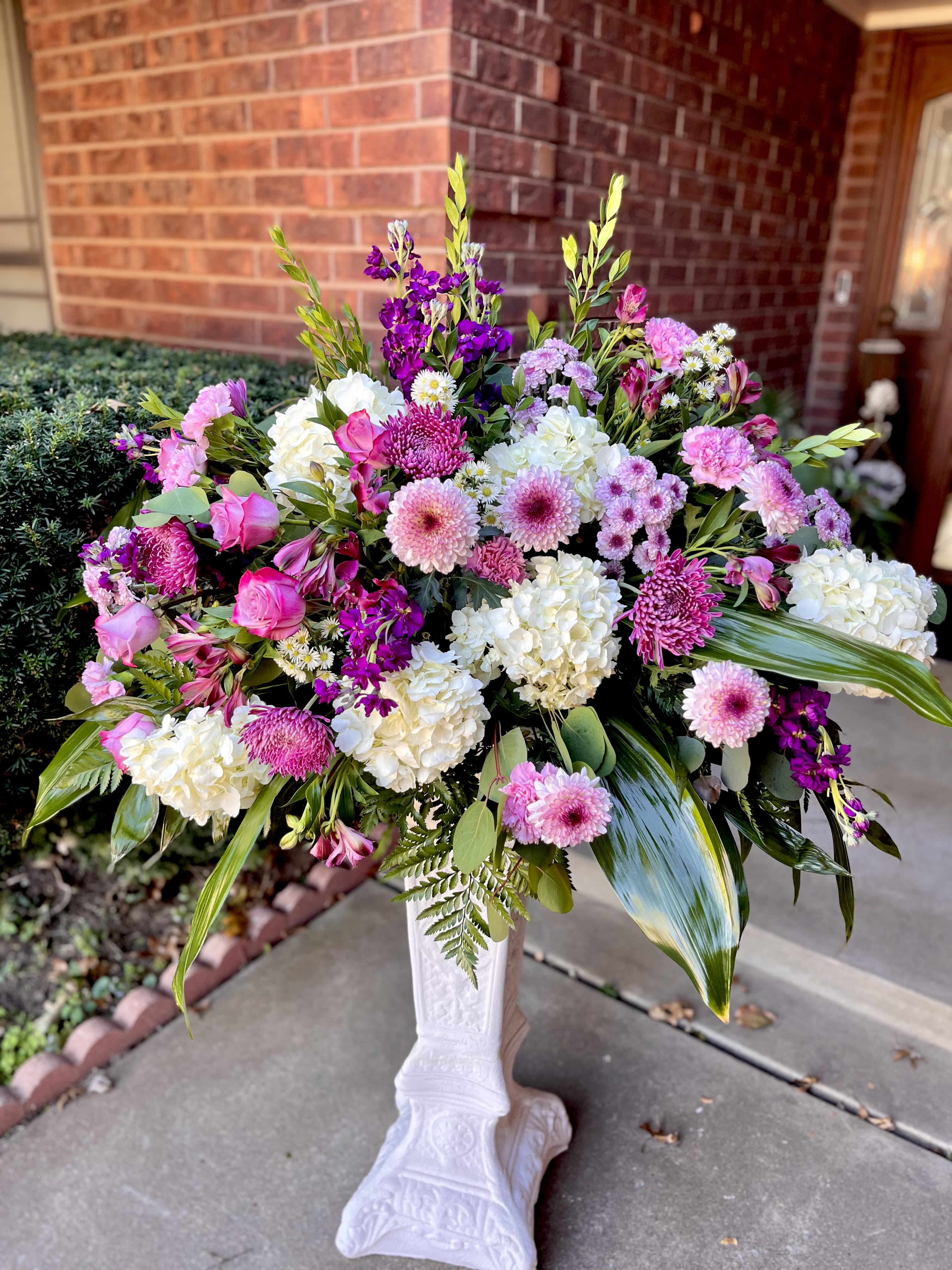 Lavender Skies - A lush, full-bodied floral arrangement built in a classic white pedestal vase, Lavender Skies is a beautiful sympathy tribute, a celebratory flower arrangement perfect for birthdays, anniversaries, or a meaningful congratulations gift. The layered clusters of pink, white, purple, and green create an immediate sense of abundance-she'll feel truly celebrated when this polished, cheerful display arrives.  This large arrangement features rounded hydrangea heads, garden-style roses, chrysanthemum and daisy-like blooms, delicate alstroemeria, and graceful orchid sprays set against broad tropical leaves for movement and contrast. The pedestal vase lifts the composition for a formal presentation and makes the colors-soft pinks, crisp whites, and cool lavenders-read beautifully from every angle.  ***Pedestal for display only