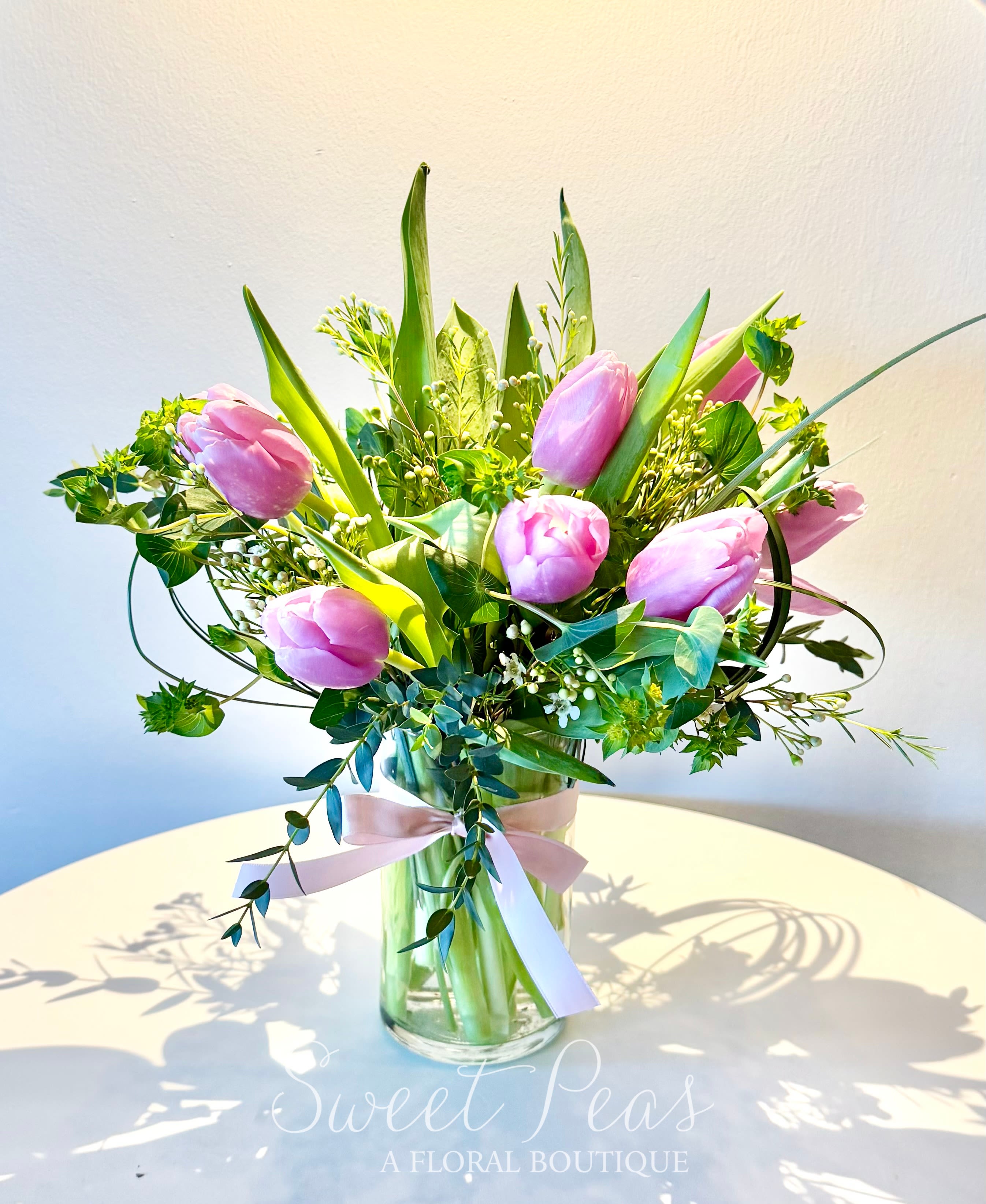 Pink tulips arranged in a clear glass vase with a pink ribbon on a white table.
