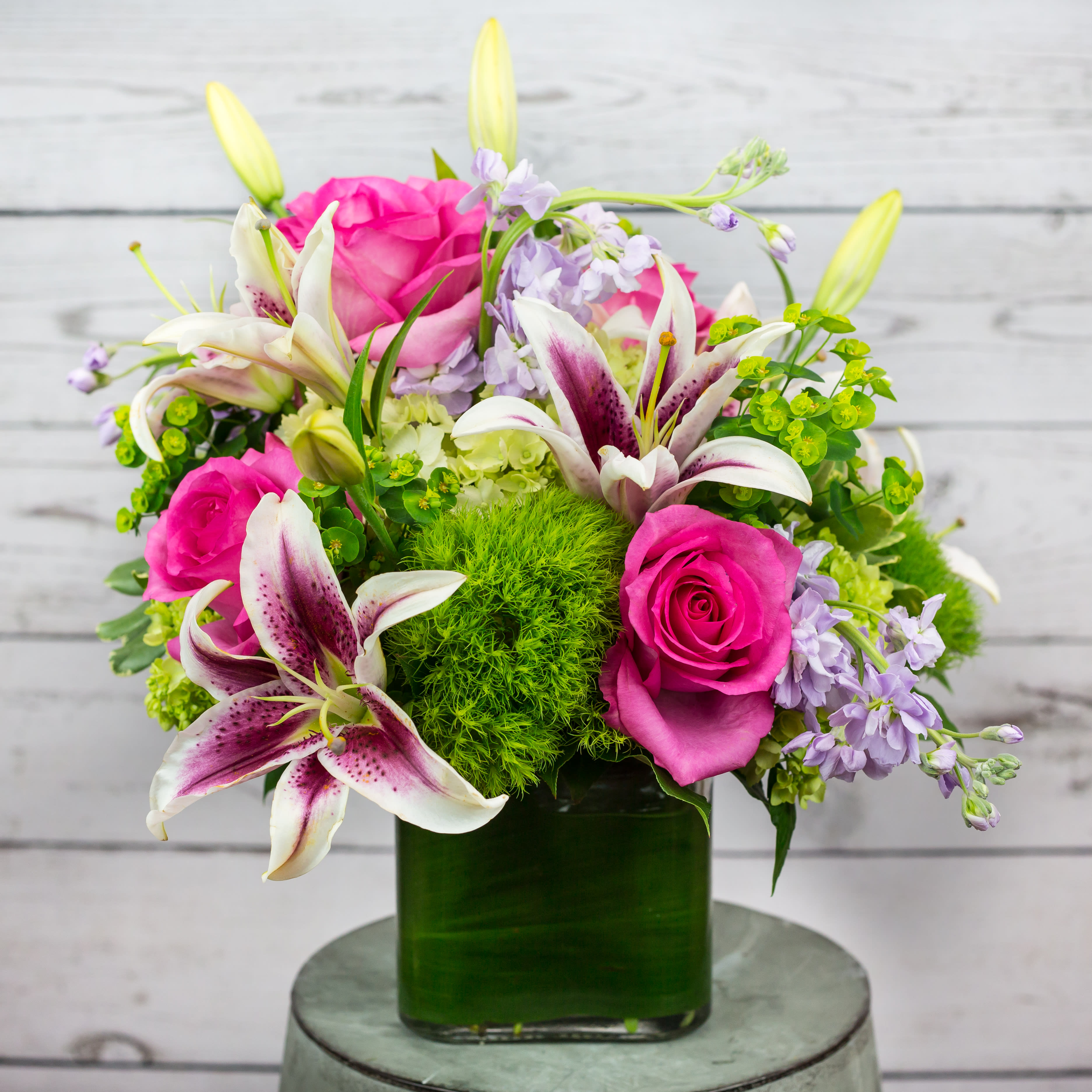 Bright arrangement of pink roses, white and magenta lilies, and lavender blooms in a green glass cube vase