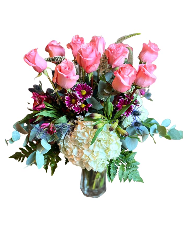 Pink Mixed DZ - Soft pink roses rise above a cloud of cream hydrangea in this pink mixed flower arrangement, a cheerful birthday bouquet or anniversary gift that reads as both romantic and playful. The rounded, abundant shape makes it the kind of floral surprise that will make the person you're thinking of feel truly celebrated - ideal for birthdays, anniversaries, or simply saying "congratulations."  This arrangement pairs velvety pink roses with pale blue eucalyptus-like foliage, purple daisy-style blooms, blue thistle-like accents, alstroemeria, chrysanthemums and other mixed seasonal stems for texture and contrast. Warm cream hydrangea forms a full base that highlights the taller rose spray; everything comes arranged in a clear glass vase for an elegant, ready-to-display presentation.  Perfect for sending to a close friend, partner, or family member who loves soft pastels and layered textures, these long-lasting blooms are arranged to show well on a dining table, mantel, or office reception. Standard sizing creates that lush, rounded silhouette pictured; order now for same‑week delivery and include a short note to personalize the gift.