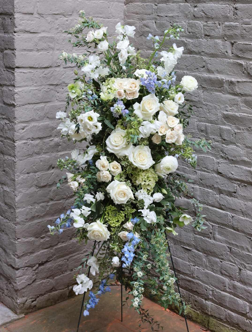 Deluxe Standing Funeral Spray with Easel - An exquisite standing display of flowers to celebrate the life of your loved one.  Our feature photo is comprised of white and blue blooms; it is our honor to accommodate any other specific color preferences, per your request. Delivered on wire easel. Measures approximately 32" long by 18" wide.  A special note: This arrangement is constructed with compostable basalt wool (in lieu of the typical floral foam and plastic). When available, we are happy to incorporate locally-farmed and harvested flowers as well. Thank you for supporting a more eco-friendly and sustainable option for funeral florals.