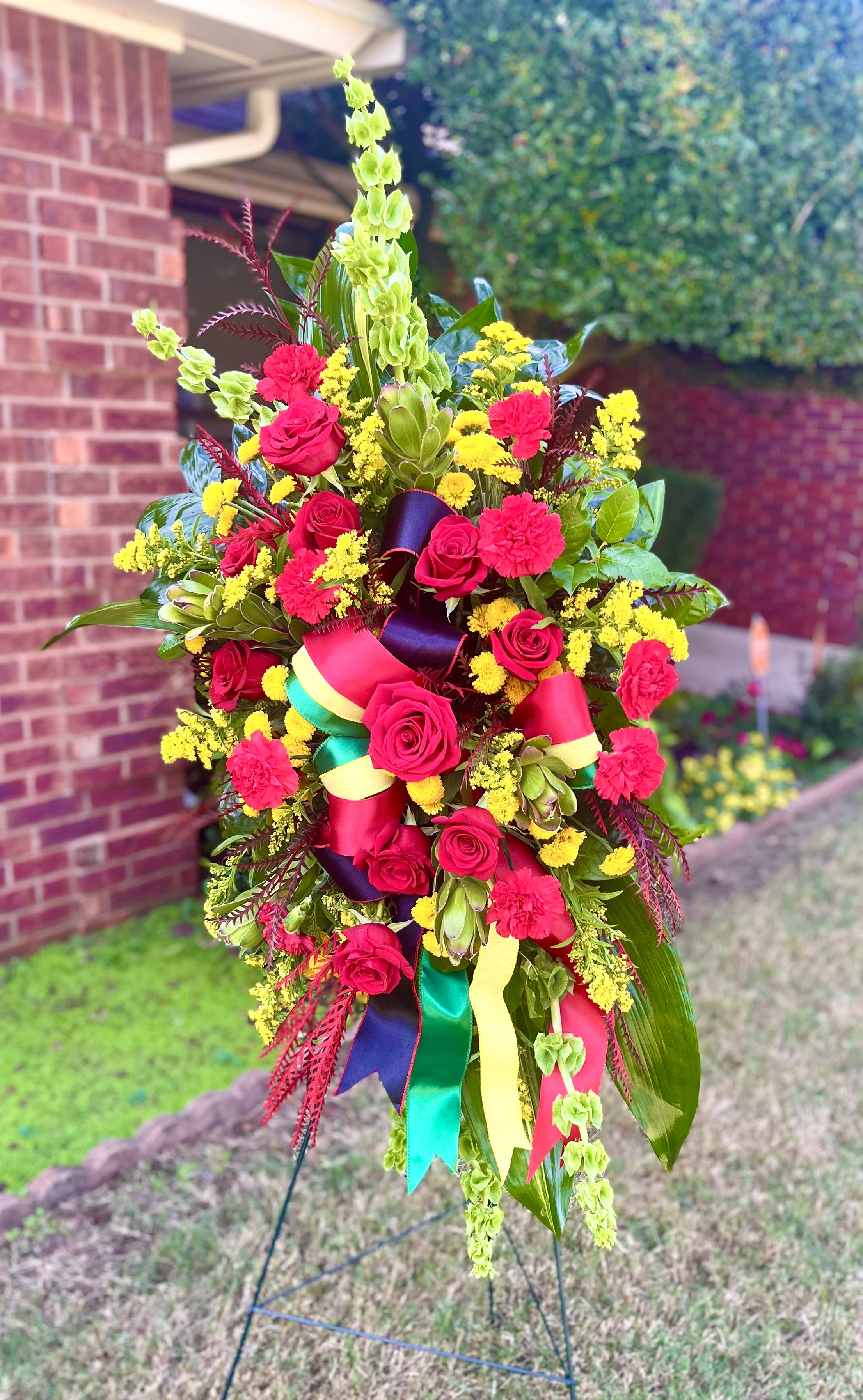 Tipton - A tall, ceremonial standing arrangement designed to make a bold statement, perfect as a sympathy tribute or for marking a special event. This funeral spray-style flower arrangement features red freedom roses and carnations as the visual center, accented with clusters of small yellow filler blooms and green blossoms that brighten the overall palette. The dramatic vertical silhouette and cascading elements ensure the recipient-or the moment-feels honored and held.  Dense, sculpted, and richly textured, the arrangement pairs long arching leaves and burgundy-toned foliage for height and contrast, while wide satin ribbons in bright colors add a festive, formal finish. Mounted on a metal easel stand, the piece is designed to be seen from a distance and photographed with dignity. Sized to command a service space or entrance, it offers the kind of presence that conveys heartfelt respect or celebratory recognition for a life, milestone, or important ceremony.