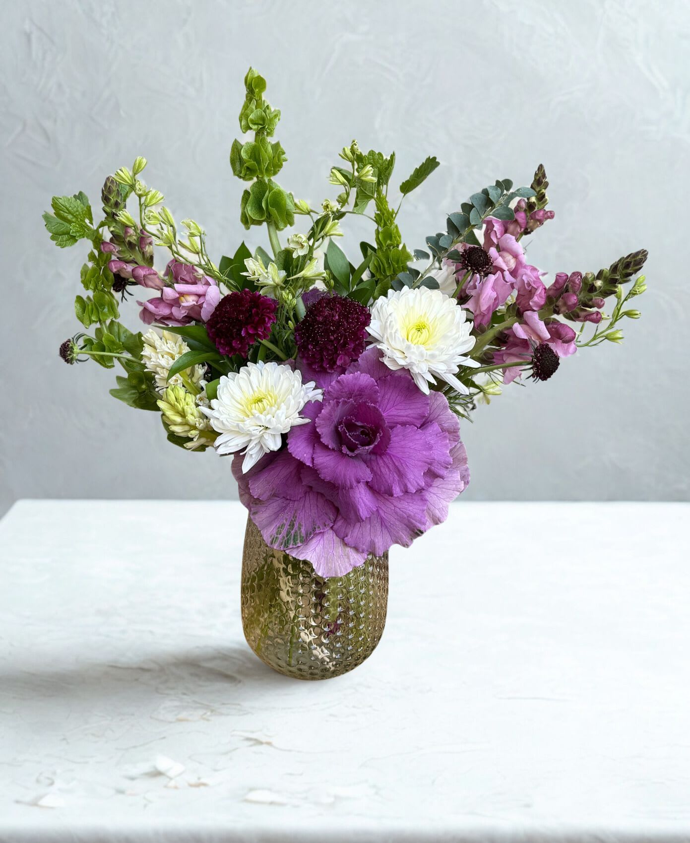 Mom's Garden Party - A fresh, garden-inspired arrangement featuring a bold purple ornamental kale, soft snapdragons, and a mix of white and deep burgundy blooms. Designed with natural movement and texture, perfect for adding seasonal charm to any space. Container color may vary.