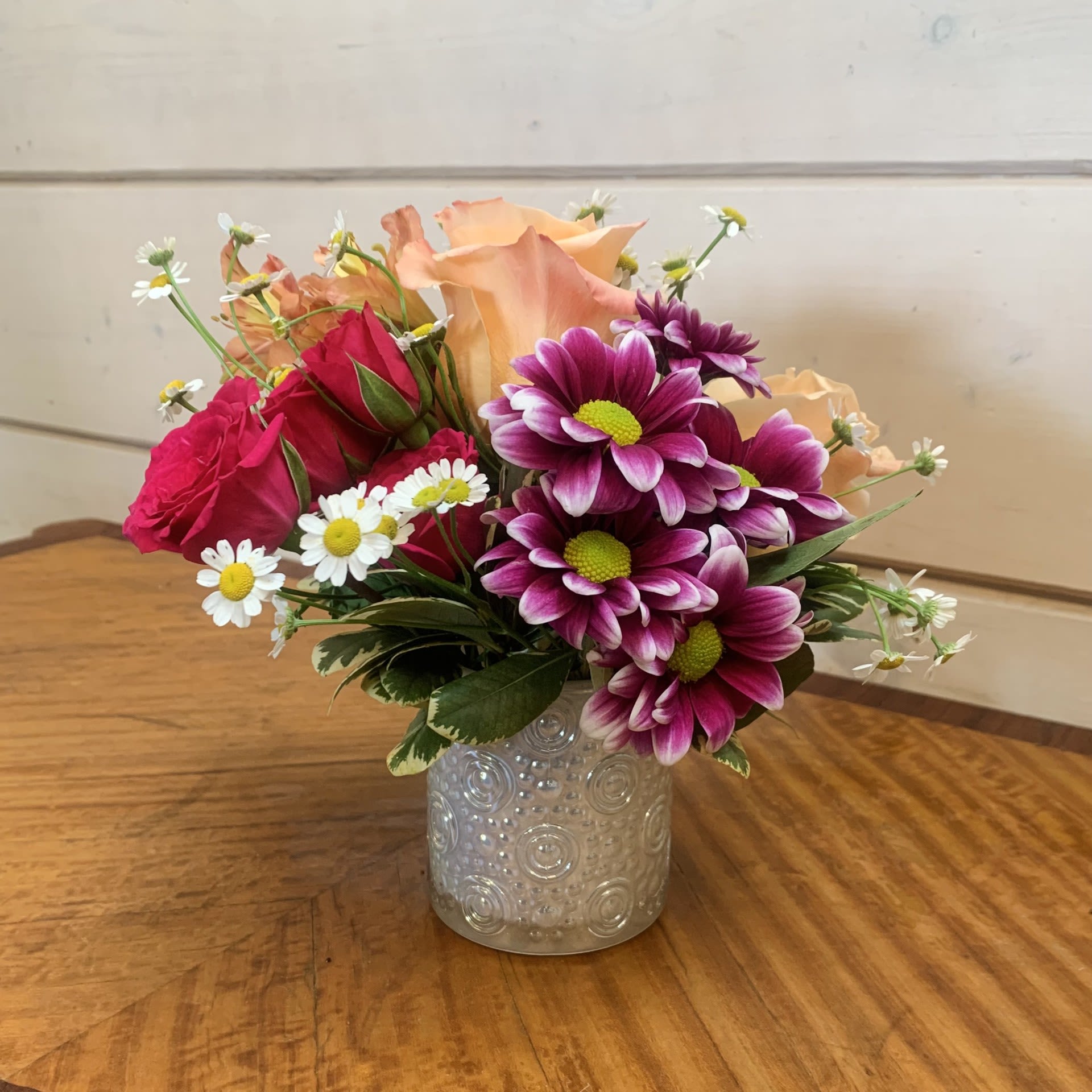 A Little Friendship  - Apricot roses and hot pink spray roses with bicolor purple daisies in a petite iridescent glass vase with orange alstroemeria and seasonal filler flower. Approximately 6" tall by 6" wide.