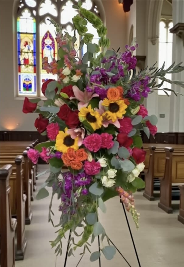 Sunset Symphony Spray - Sunset Symphony is a bold Celebration of Life standing spray designed to honor a spirit that lived fully and brightly. Featuring radiant sunflowers, rich red roses, hot pink carnations, coral spray roses, lilies, and vibrant seasonal blooms, this design is layered with color, texture, and movement from top to base.  Strong vertical lines and a natural cascade of eucalyptus and greenery give the piece height, flow, and presence, creating a striking silhouette that stands out beautifully during the service.  The color story is warm, energetic, and expressive—designed to reflect a life filled with personality, love, and unforgettable moments.  A powerful choice for families who want something uplifting, vibrant, and full of life—without losing the sense of respect and intention the moment deserves.  Handcrafted by Aspen Tower Florist and available for funeral flower delivery in Greensburg, Indiana and surrounding areas including Shelbyville, Rushville, Batesville, Westport, Oldenburg, St. Paul, Waldron, Milroy, and Hope. 