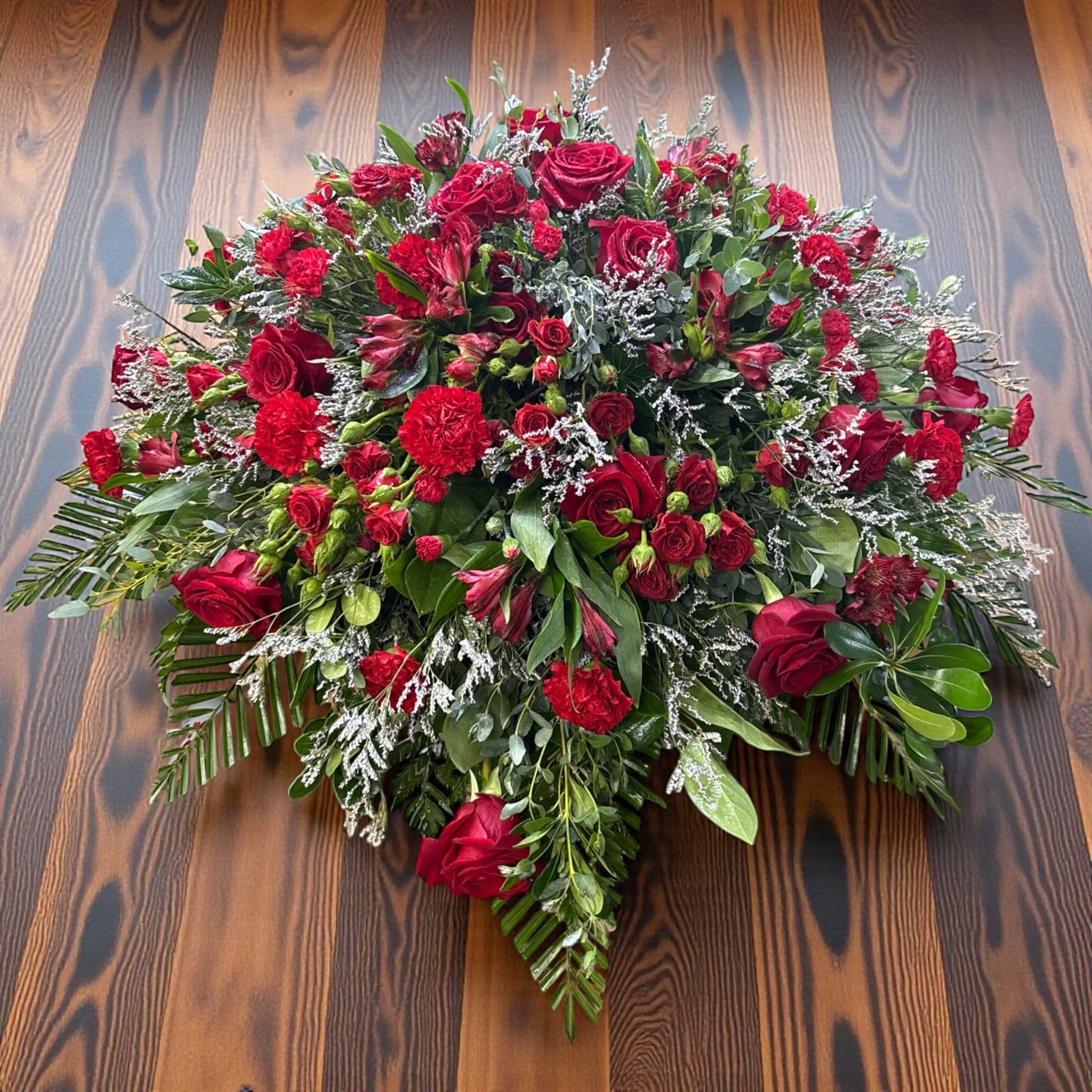 Tribute of Life - Tribute of Life  Tribute of Life is a bold, all-red casket spray designed to make a powerful, unified statement of love and remembrance. Featuring red roses, spray roses, carnations, mini carnations, alstroemeria, and accents of limonium, this design creates a rich, layered field of color with depth, texture, and movement throughout.  The strength of this piece comes from its consistency—every bloom working together in a single, uninterrupted palette of red, symbolizing deep love, respect, and enduring devotion.  Clean lines, balanced structure, and full coverage give this spray a strong visual presence across the casket, allowing it to read beautifully from every angle during visitation and services.  This design gained widespread attention after a creation video went viral on Pinterest, resonating with thousands for its striking simplicity and craftsmanship.  A meaningful choice for families who want something timeless, powerful, and deeply heartfelt.  Handcrafted by Aspen Tower Florist and available for funeral flower delivery in Greensburg, Indiana and surrounding areas including Shelbyville, Rushville, Batesville, Westport, Oldenburg, St. Paul, Waldron, Milroy, and Hope.