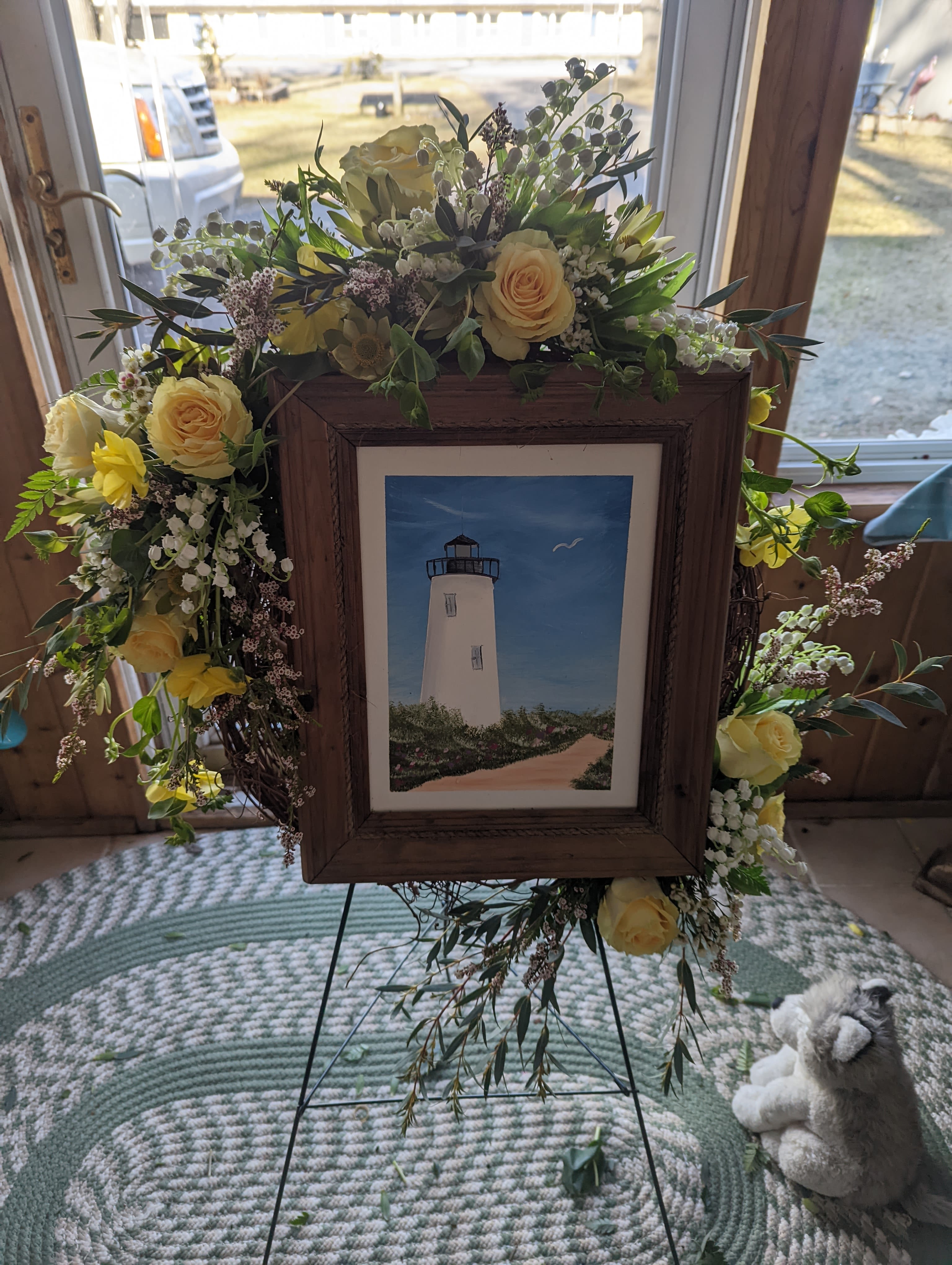 Funeral Wreath with Picture - A framed lighthouse painting rests at the heart of this sympathy wreath, a rustic memorial arrangement that reads as a gentle, sea‑kissed tribute for a funeral or remembrance service. The wooden picture frame is embraced by a loose garland of pale yellow roses, white daisy‑style blooms, soft pink accents, and airy green foliage, creating a calm, comforting presence that will help the person you're honoring feel remembered and seen.  Flowers include pale yellow roses, white daisies, small filler blossoms, and mixed greenery arranged to drape naturally around the frame; a small stuffed animal can be included for a child's memorial or to add a tender touch. The wreath is displayed on a metal easel for easy placement at a service or graveside, and its approachable, homey texture lends itself to heartfelt remembrances rather than formal funeral displays.