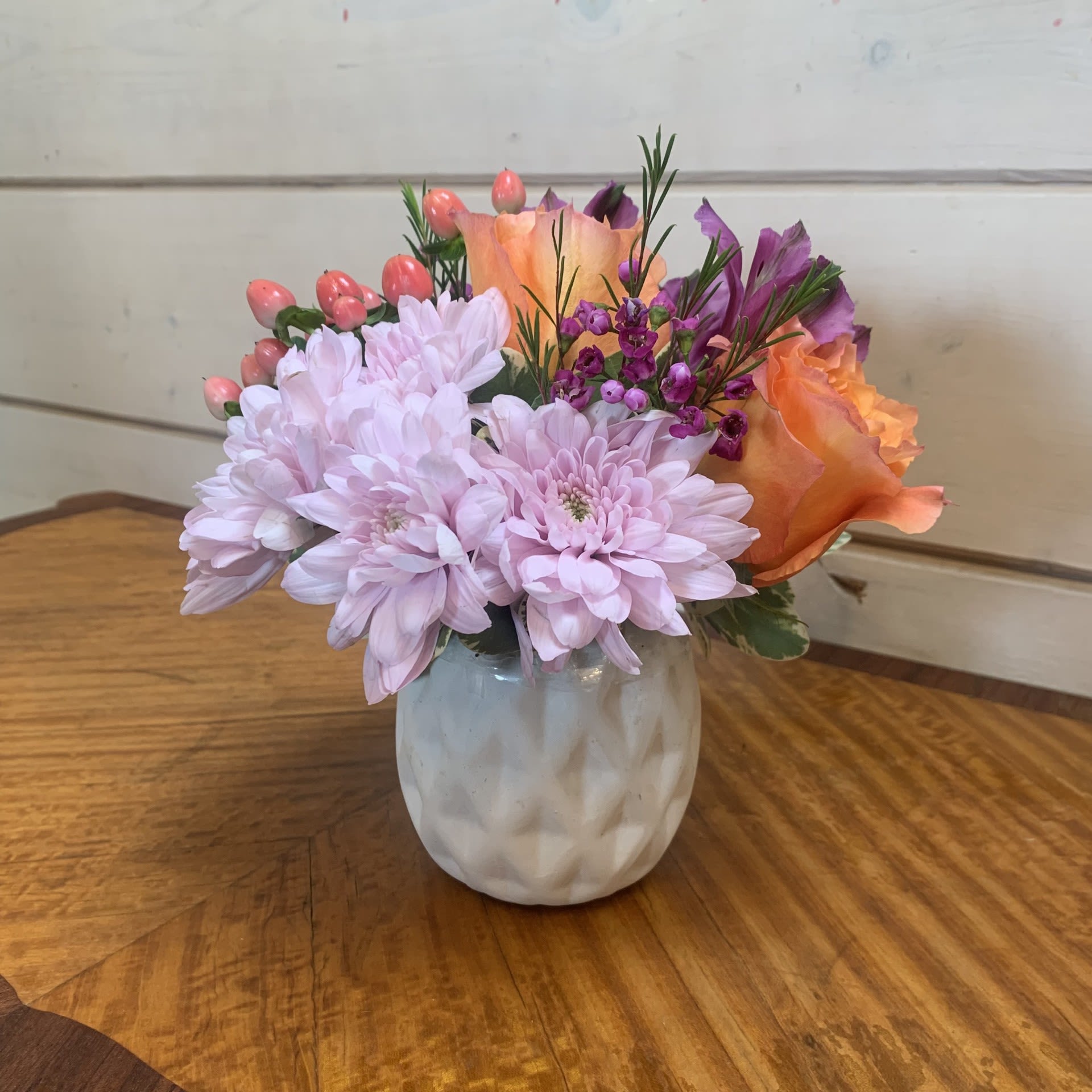 A Little Spring - A petite arrangement of apricot roses, pink hypericum berries, and pink cushion chrysanthemums in a white patterned pot with purple alstroemeria and seasonal filler flower. Approximately 6" tall and 6" wide.