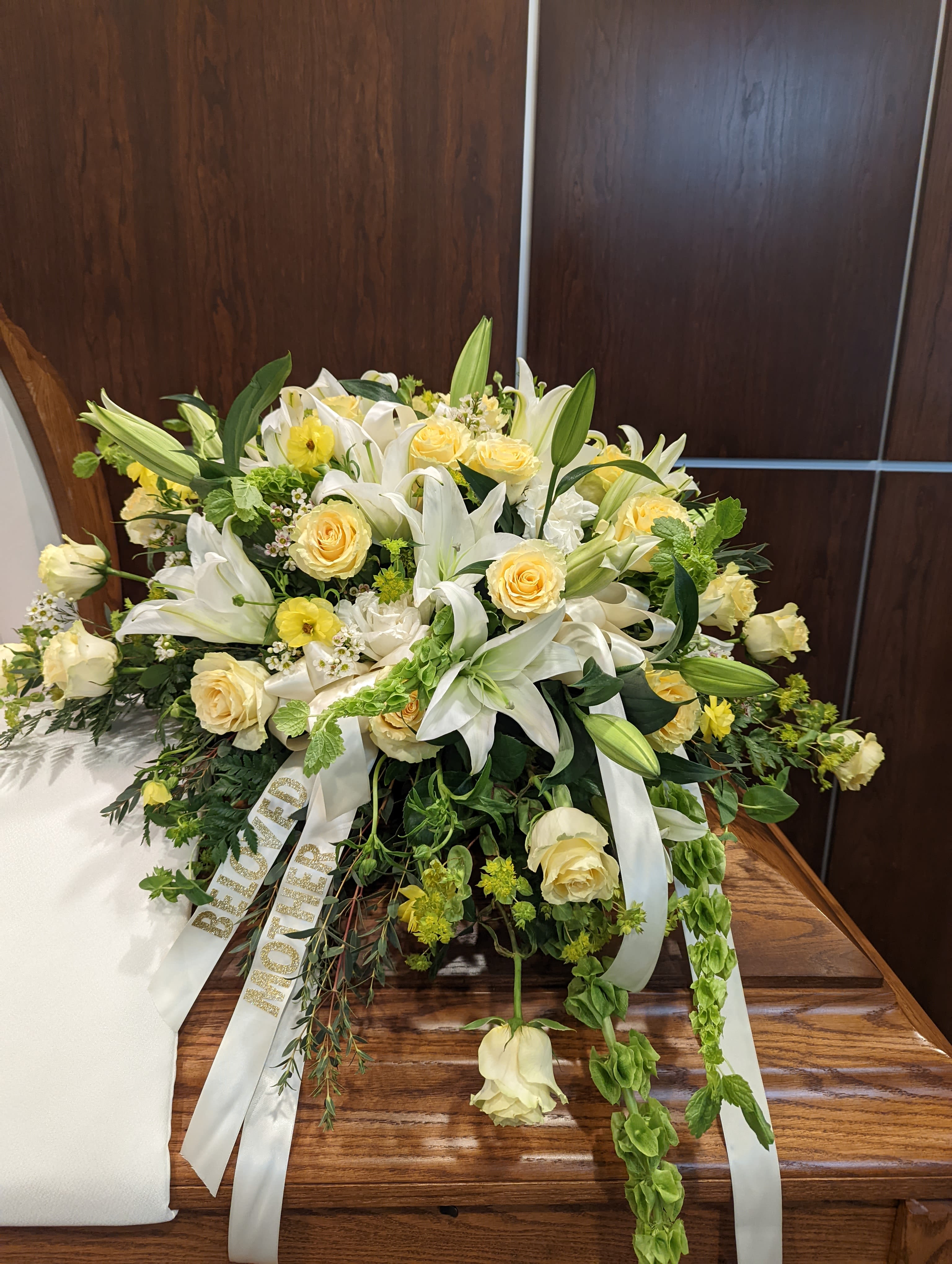 Funeral Casket Spray - A graceful, full-length casket spray designed for a funeral or memorial service, this arrangement combines white lilies and pale yellow roses to convey sympathy and quiet warmth. The soft ivory-and-lemon palette and cascading greenery create a serene, comforting presence at a service-an arrangement meant to help the person you're honoring feel remembered and cherished.  Built low and lush, the spray mixes open lilies with lily buds, pale yellow roses, and small accent blooms for depth and texture, finished with trailing greenery for a layered, flowing silhouette. White satin ribbons with glittered lettering hang from the front to give the piece a formal, ceremonial touch; the substantial size is crafted to rest across a casket or memorial display with dignity.  Appropriate for a traditional funeral or celebration of life, this spray offers a respectful way to express condolence to family and friends. Care is simple: keep the foam moist until placement and avoid direct heat; if you need a specific size or personalization on the ribbon, please contact us when ordering so we can ensure it fits the service.