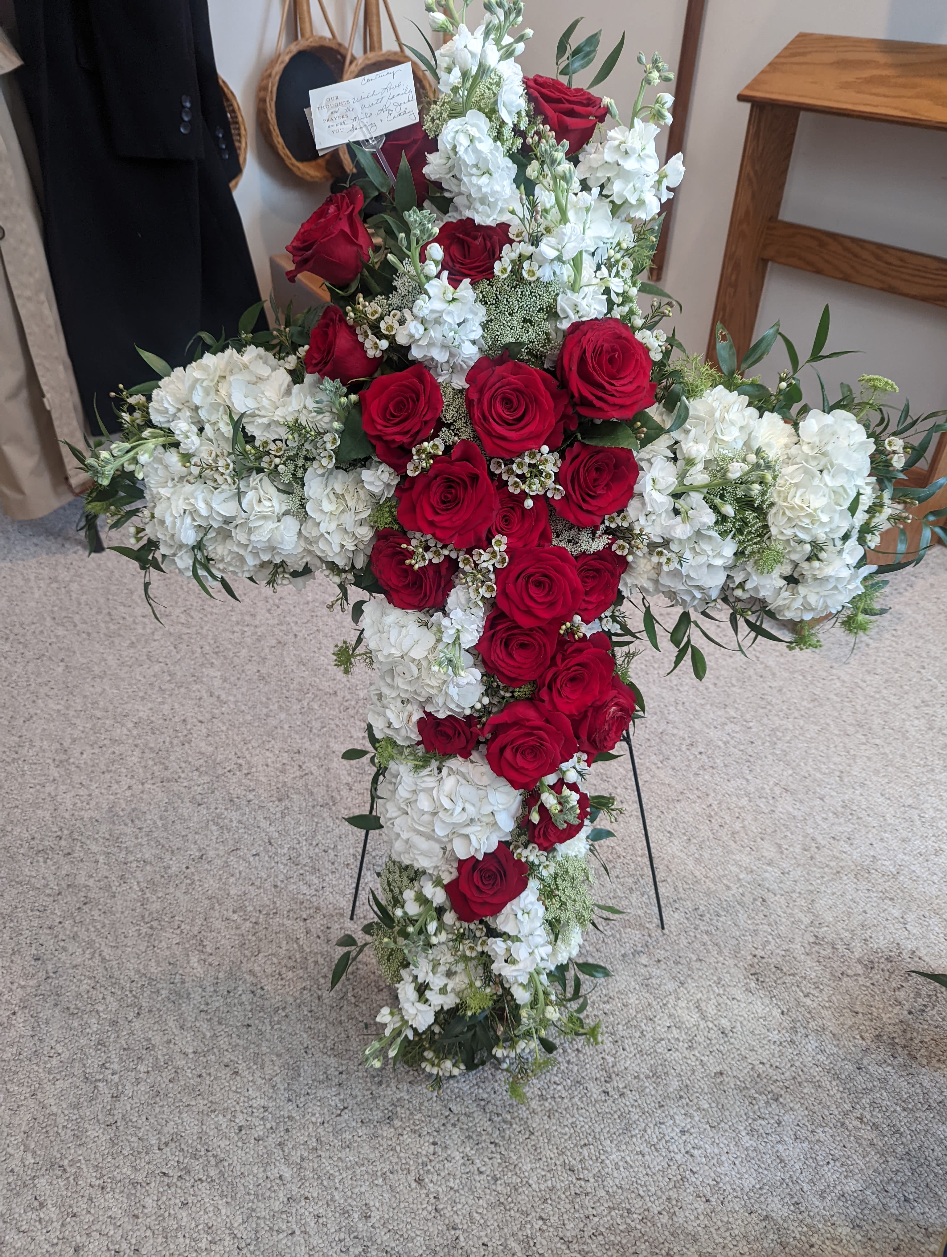 Funeral Cross - A large, traditional sympathy cross arranged for a funeral service, this floral tribute centers on deep red roses laid into the heart of the cross with white hydrangea-like blooms and tall white stock creating a soft, reverent border. The red, white, and green palette reads formal and comforting, designed to convey respect and lasting remembrance for the person you're honoring.  Dense clusters of roses form the bold central field while the white blooms and upright stock add height and an elegant, serene contrast; small filler flowers and airy greenery soften the edges and keep the cross feeling full and balanced. The arrangement is finished for display on a metal easel and includes a card holder for personalization, making it ready for placement at a service or graveside.