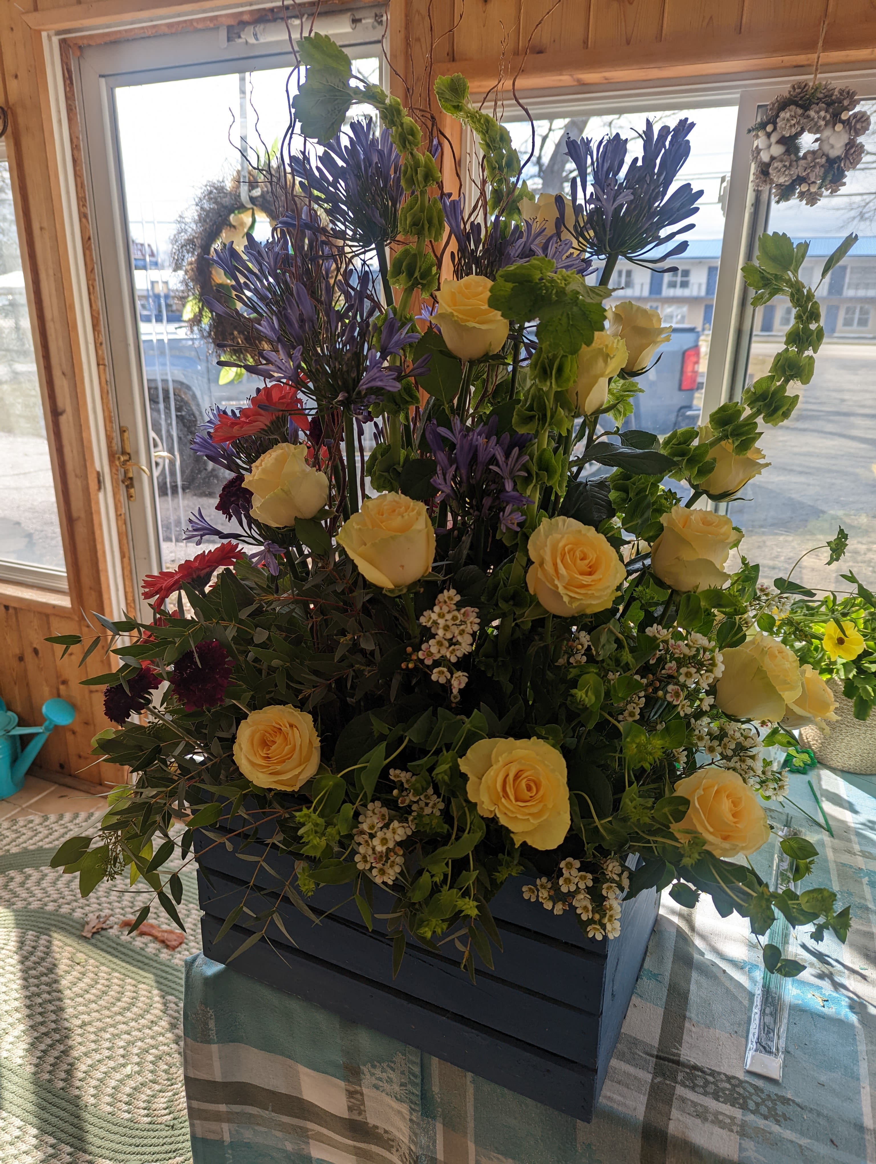 Funeral urn - Pale yellow roses take center stage in this tall, layered funeral urn arrangement, offering a soft, comforting focal point that reads both celebratory and respectfully formal for a memorial service or sympathy display. The upright, garden-like silhouette combines height and abundance so the person you're honoring will be framed by gentle color and texture-an arrangement suited to a funeral service, memorial table, or bedside tribute.  Purple spiky blooms and small white filler flowers weave through the roses to add vertical contrast and delicate texture, while a few red daisy-like blossoms bring warm accents that brighten the composition without overpowering it. Designed in a dark rectangular wooden container, the piece feels grounded and structured; its full, layered form and mixed bouquet styling create a lasting visual presence appropriate for formal settings.  This arrangement is intended to convey comfort and thoughtful remembrance-the kind of floral tribute that helps mourners feel seen and supported. The sturdy wooden container makes it easy to place on a casket spray area, memorial table, or near a service entrance; standard care is to keep the flowers in fresh water and away from direct heat to preserve their shape and color during services.