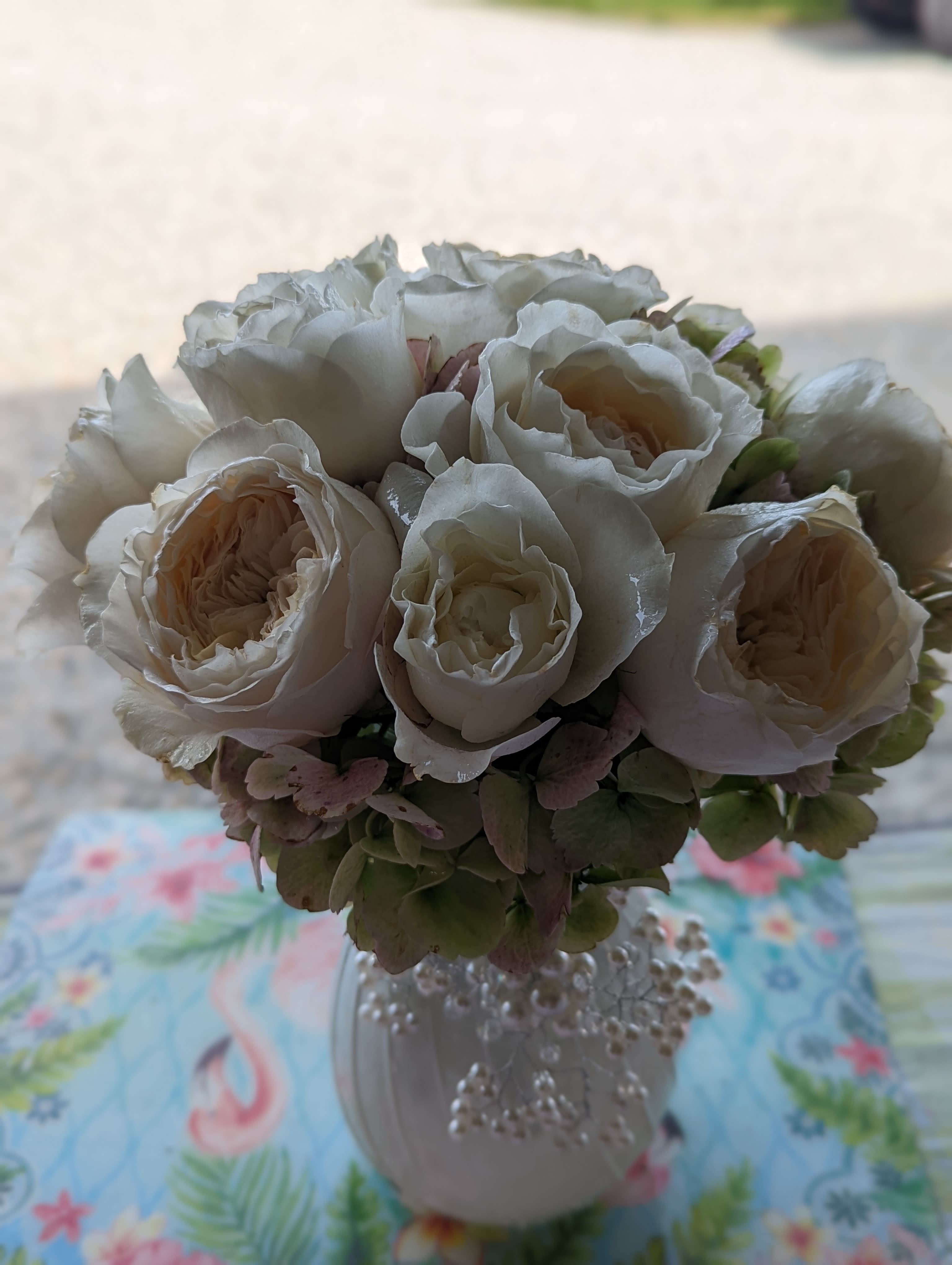 Sweetheart Vase - Creamy white roses gathered into a compact, rounded bouquet create an immediate sense of gentle romance in this flower arrangement-perfect as a wedding centerpiece or an anniversary bouquet that says "you're cherished." The soft pink-tinted hydrangeas nestle around the roses, adding a muted pastel contrast and a cloud-like texture that makes the recipient feel thoughtfully celebrated.  Arranged in a small white ceramic vase, the design is finished with delicate pearl-like strands that drape down for an elegant, feminine touch. Colors are a soothing palette of white, pink, and green; the roses provide classic form while the hydrangeas give fullness and movement. The vase's compact size suits tabletops, bedside stands, or office desks without overwhelming the space.  Ideal for brides, mothers, or anyone who loves refined, romantic florals, this arrangement reads as both a wedding accent and a lovely everyday gift. Order with confidence for same-week celebrations or to send a thoughtful surprise; simple care-fresh water and a cool spot away from direct sun-will keep the blooms looking their best for days.