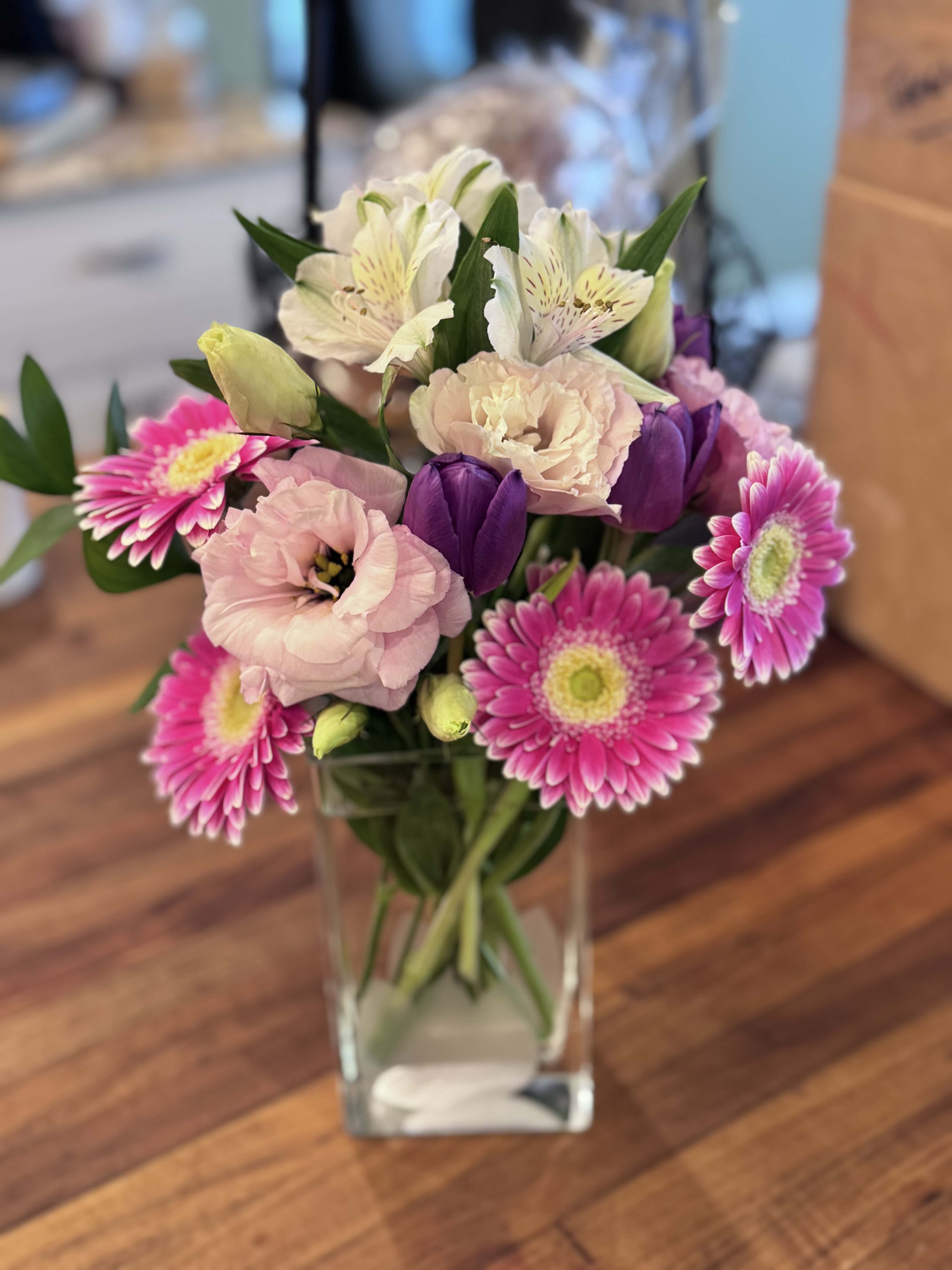 Spring vase - A tall, airy spring vase that reads like a fresh garden in bloom-perfect as a birthday bouquet, a thoughtful thank-you, or a sympathy-style arrangement that comforts. Pink roses and pale lilies give a gentle, romantic focus while pops of purple tulips and bright gerbera daisies lift the mood; the arrangement's vertical, loose composition feels elegant and slightly formal so the person you're sending it to will feel truly seen and comforted.  This design is layered with soft blush lisianthus, white alstroemeria, alstroemeria and mixed fine filler blooms tucked into abundant greenery for texture and movement. Depending on the option shown, it is presented either in a white ceramic vase with blue stripes on a small display stand (with a small card tucked into the stems) for a refined, garden-like statement, or in a tall clear glass vase that highlights the stems and creates a lively, cheerful display. Choose the vase that best suits the occasion; stems arrive arranged and ready to place, requiring only fresh water to keep them looking their best.