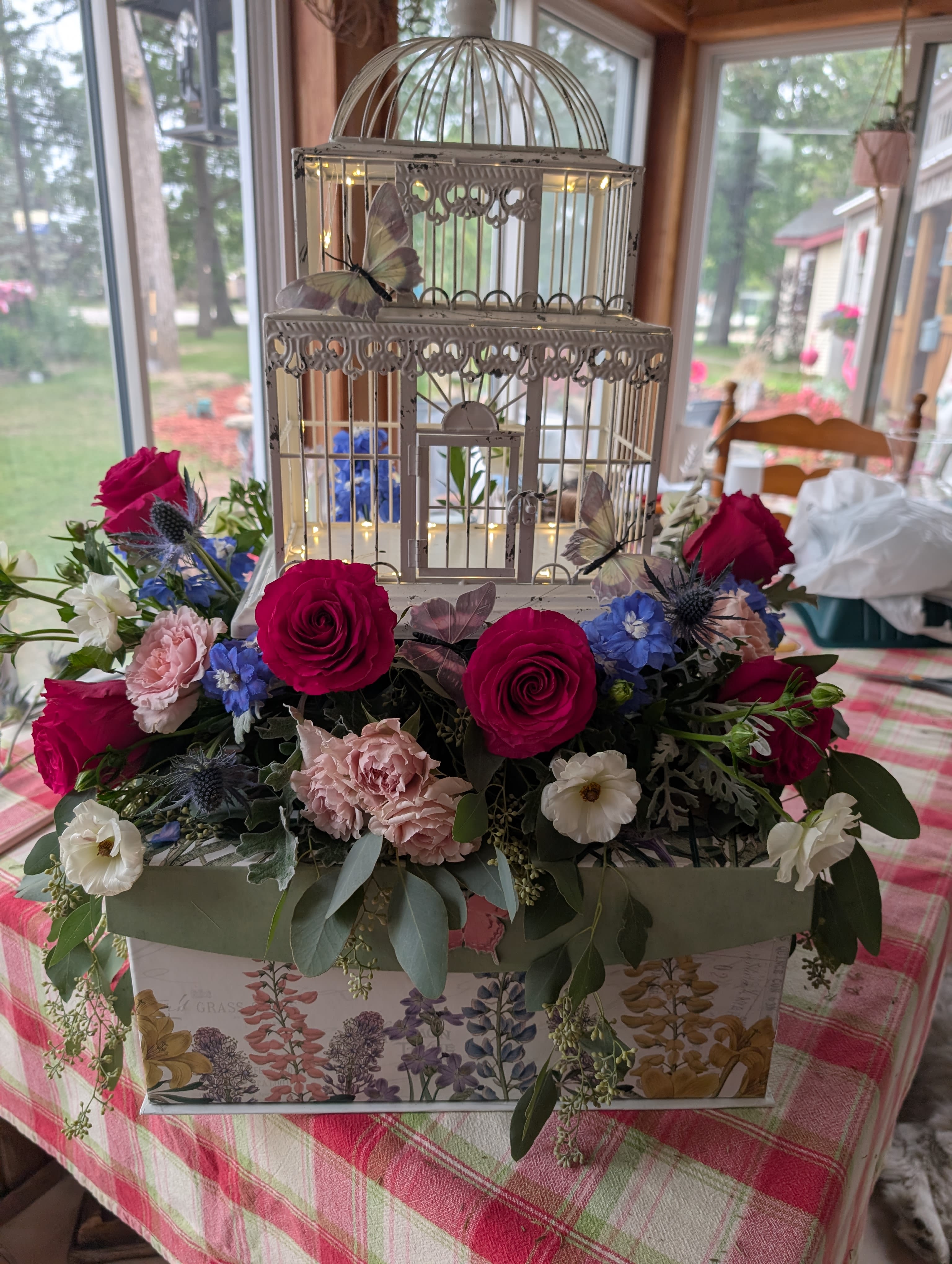 Bird cage on a Box - A whimsical white birdcage nestles into a decorative box framed by a dense ring of bright pink roses, making this floral arrangement a charming centerpiece for a birthday table, spring celebration, or romantic anniversary. The hand-crafted look-with butterfly ornaments perched among the blooms-reads playful and romantic; the person you're sending it to will feel delighted and noticed the moment it arrives.  Soft pale-pink carnations and small white blossoms layer around the vivid roses, while touches of blue flowers and fresh greenery add cool contrast and depth. The mix of roses, carnations, daisies, and other seasonal blooms creates a textured, lively bouquet that looks full from every angle. The decorative birdcage and box give the piece structure and make it easy to display on a dining table, mantel, or entryway.  Ideal for someone who loves whimsical, garden-inspired decor, this piece works beautifully for birthdays, Mother's Day, engagements, or a cheerful "thinking of you" surprise. Arrange on a flat surface and keep blooms away from direct heat; fresh water and occasional misting will help the flowers stay vibrant during the celebration.