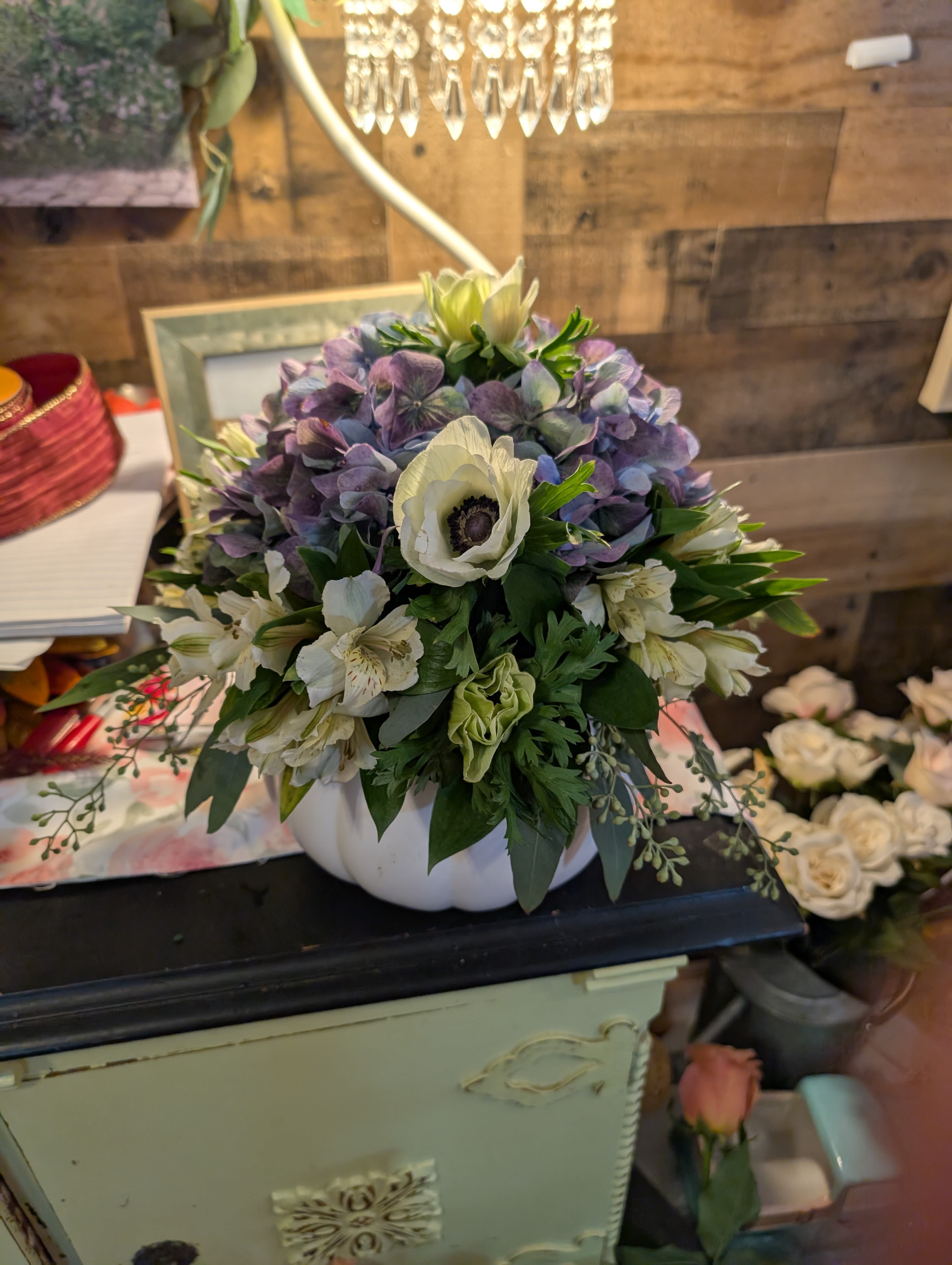 White vase - A rounded mound of purple hydrangeas forms the soft, fragrant heart of this fall tabletop arrangement, accented by cream-white anemones and delicate white alstroemeria-an ideal flower arrangement for autumn gifts or a seasonal centerpiece. The compact bouquet sits snugly in a white ceramic pumpkin vase, so the overall presentation reads cozy and decorative while still feeling refined for a birthday, hostess gift, or Thanksgiving table.  The palette blends soft lavender, crisp white, and fresh green foliage for a calm, full look; hydrangea heads provide lush volume, anemones add contrast with their dark centers, and alstroemeria brings light, airy texture around the edges. The ceramic pumpkin vessel is durable and display-ready, making this arrangement an easy way to brighten a mantel, dining table, or office without extra staging.  Thoughtful for the person you're celebrating-this is the kind of arrangement that makes someone feel noticed and comforted during autumn gatherings or simple everyday moments. Sized as a compact centerpiece, it arrives arranged and ready to display; enjoy the bloom-rich silhouette indoors away from direct heat to keep flowers looking fresh longer.