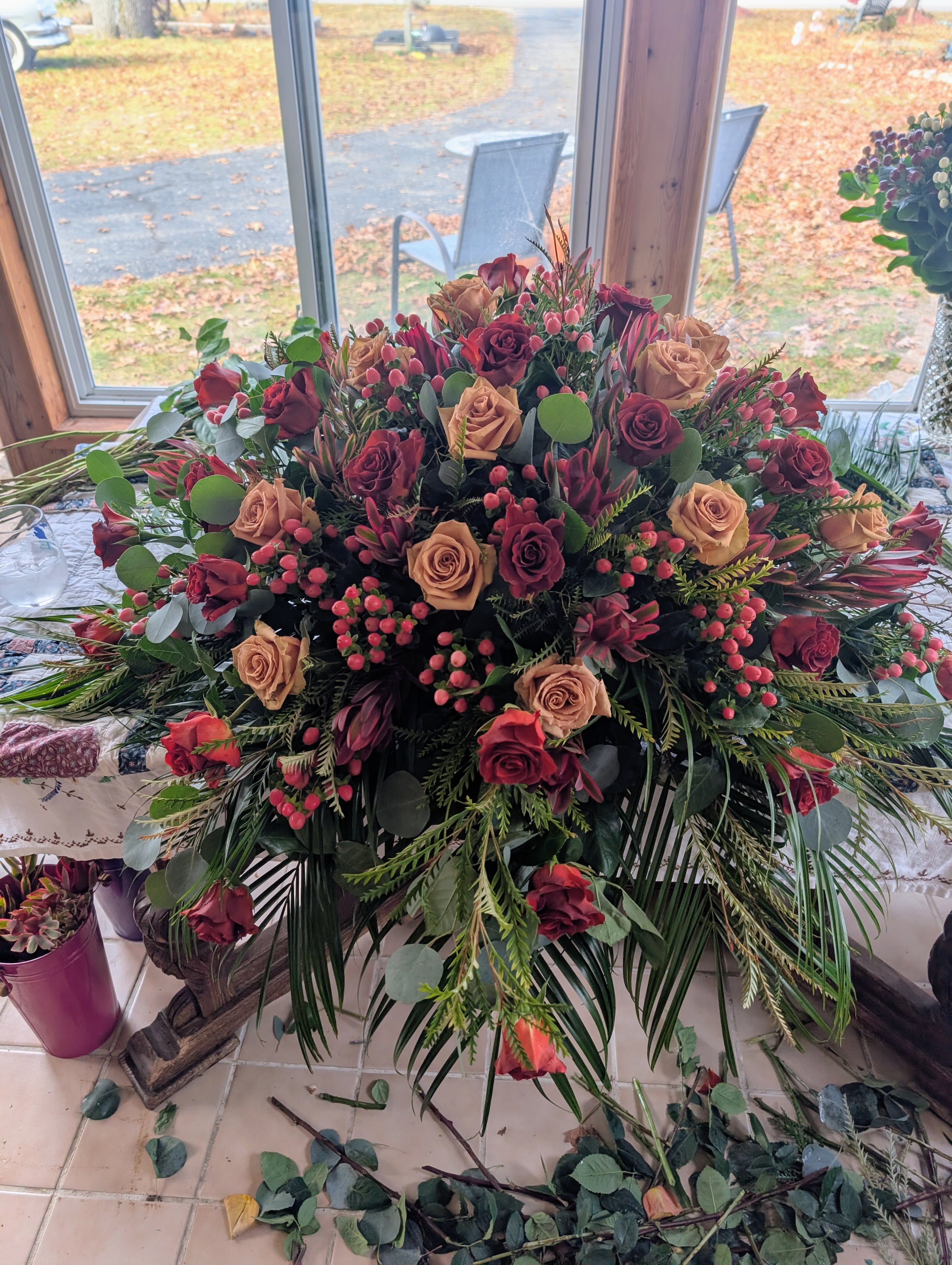 Fall Casket Spray - A lush, low centerpiece arranged for maximum table impact, this sympathy-style floral display pairs burgundy and peach roses to create a rich, romantic mood perfect for sympathy, memorial services, or a formal anniversary table. The wide, spreading design and layered textures make it a show-stopping focal point that lets the person you're honoring feel remembered with depth and warmth.  Peach and deep red roses are nestled among clusters of pink berries and mixed greenery, with long, arching foliage that adds graceful lines across the low container. The arrangement sits broadly and densely in a shallow vessel, creating a finished, polished silhouette from every angle; the contrast between the velvety burgundy and the soft peach brings both drama and tenderness to the composition.  Choose this piece when you want to convey heartfelt sympathy, profound respect, or romantic reverence-the kind of arrangement that makes the recipient feel genuinely seen and comforted. Designed to sit low on a table, it's ready to display on a mantel, reception table, or beside a casket; water and simple care for the roses and greens will keep the arrangement looking fresh through the occasion.