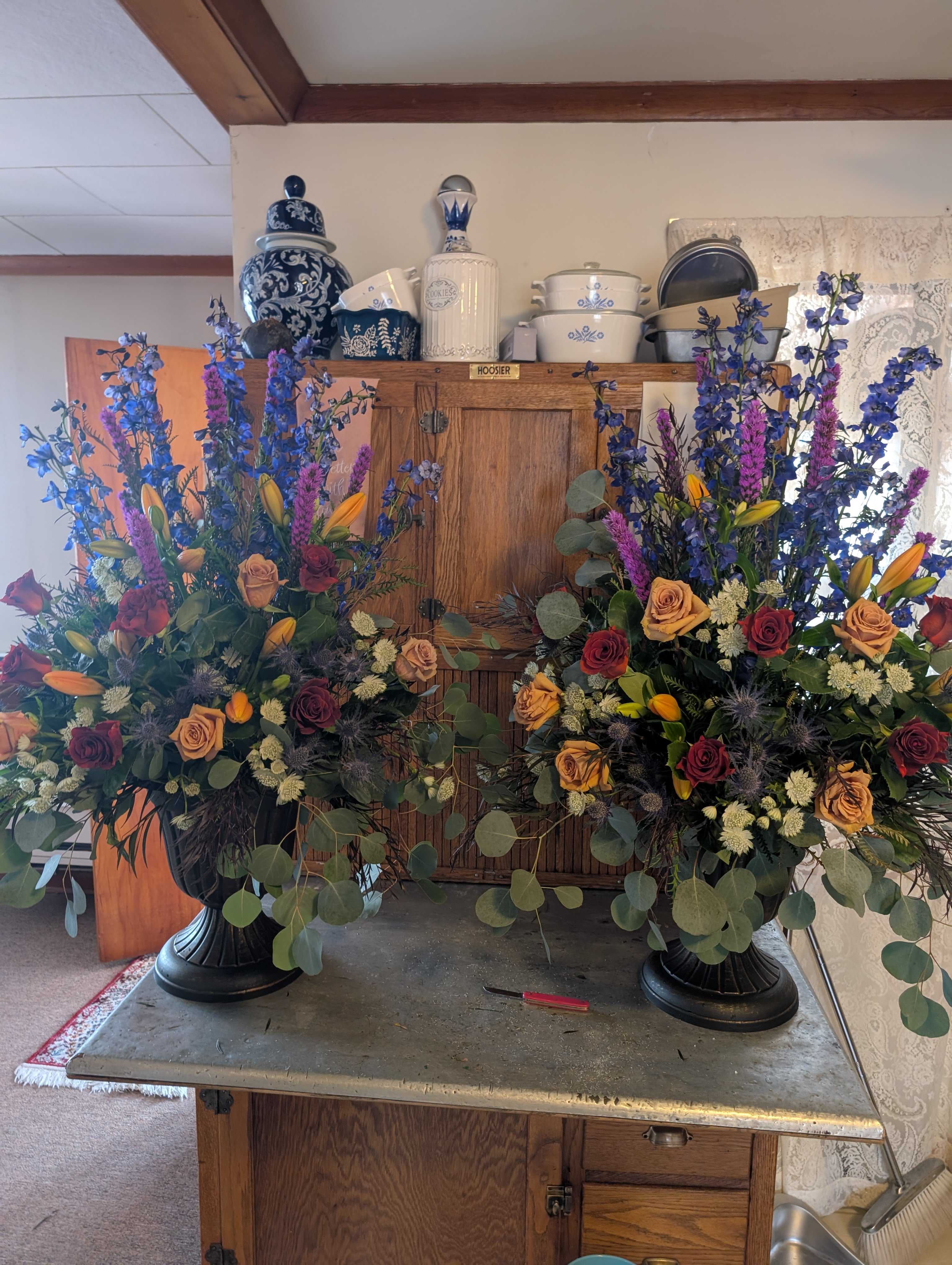 Funeral Side Sprays - $250 each - two matching side sprays shown (price is per arrangement). These formal funeral side sprays bring a rich, ceremonial presence to a memorial or celebration of life, sending a clear message of respect and remembrance. The tall, structured profile and dramatic color mix make each spray especially suited for a funeral service, chapel display, or as matching pieces beside a casket.  Each spray is arranged in a black urn vase and features deep red and peach roses paired with tall blue delphinium, purple spike flowers, white filler blooms, orange lily buds, and cascading eucalyptus for movement and fullness. The combination of vertical lines from the delphinium and lilies with layered roses creates a textured, elegant silhouette roughly matching the scale shown in the photo; colors read as rich blues, purples, pinks, and crisp white accents against the dark urn.  These side sprays are meant for a recipient or service where a formal, dignified floral tribute is desired-ideal for honoring a loved one or coordinating with other arrangements. Care: placed in their urns with floral foam, they require minimal maintenance; keep in a cool place out of direct sun until the service.