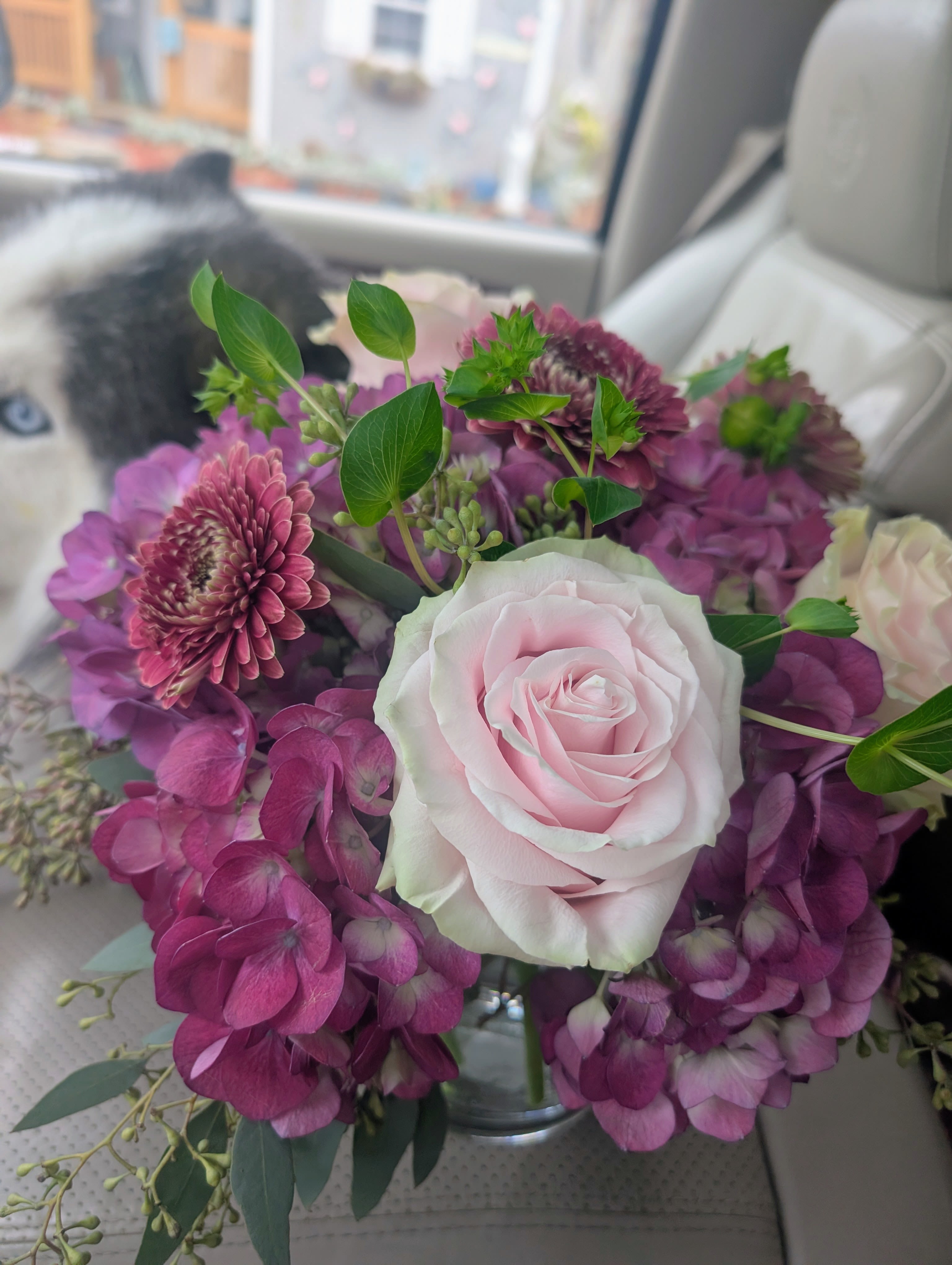 Cube arrangement - A pale pink rose takes center stage in this cube arrangement, its soft petals set against clusters of deep magenta and lavender hydrangeas for a romantic birthday bouquet or anniversary arrangement that reads both elegant and joyful. The mix of pink, purple, white, and fresh green tones creates a cheerful, heartfelt message-perfect for making the person you're thinking of feel truly celebrated on a special day or simply as a thoughtful surprise.  Surrounding the focal rose, small purple daisy-like blooms add delicate texture while airy green foliage lifts the composition into a loose, garden-style feel. The florals arrive arranged in a clear glass vase, the compact cube shape giving the display a modern, polished silhouette that still feels hand-tied and natural. This piece suits birthdays, anniversaries, or "just because" moments when you want the recipient to feel seen and adored. Care: keep water fresh and trim stems every few days to extend bloom life.