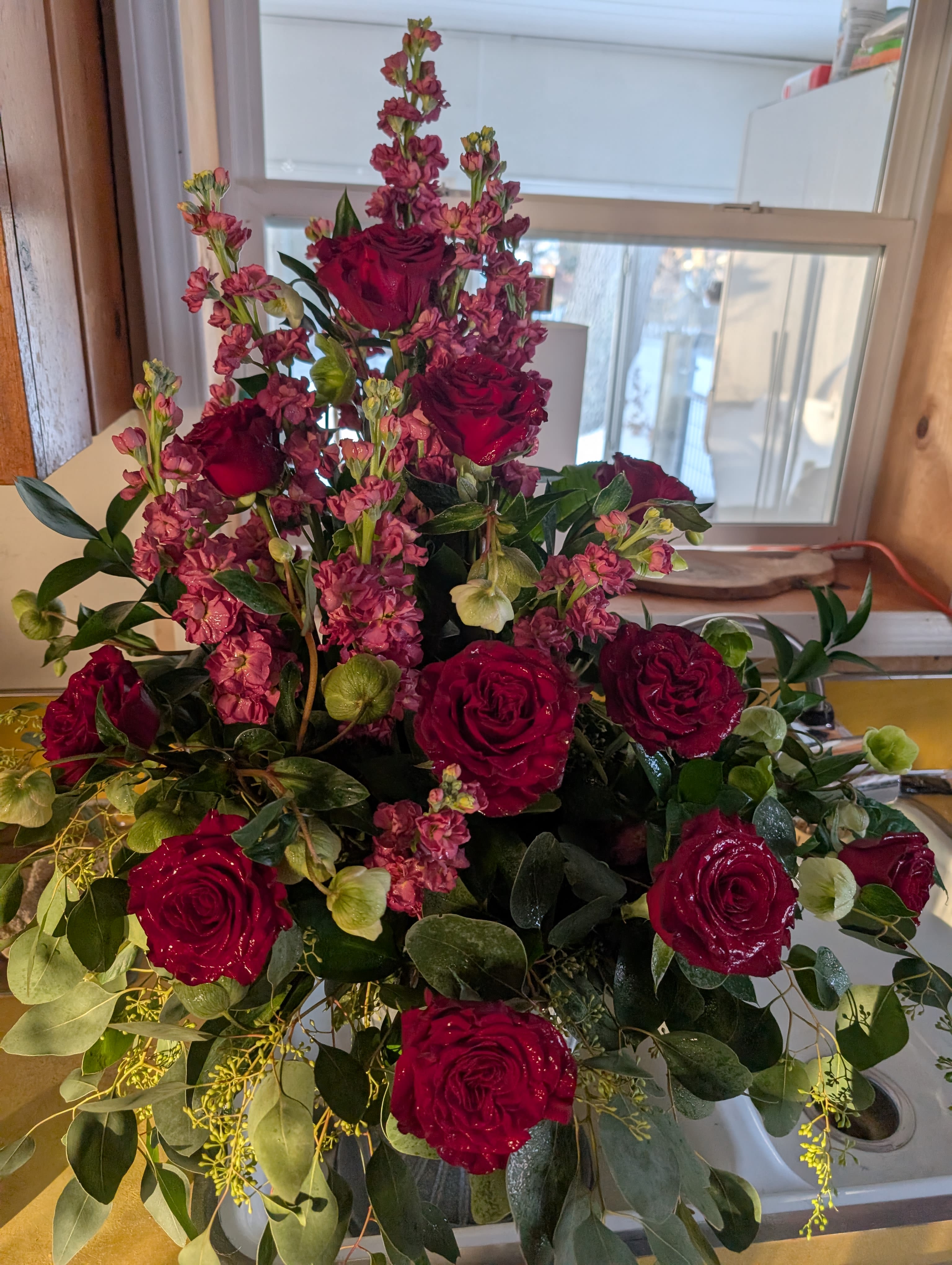 Funeral Spray - A lush, formal funeral spray that reads with quiet romance-deep red roses clustered against tall stems of soft pink stock create a vertical, layered composition ideal for a sympathy setting or a memorial service. This funeral arrangement sends a message of heartfelt love and remembrance, the kind that comforts the person you're thinking of and honors a life with dignity.  The design pairs richly hued red roses with delicate pink stock and abundant green foliage, finished with touches of white to lift the palette. The vertical silhouette gives the spray a substantial, elegant presence on an easel or casket end; textures move from the velvety rose heads to the feathery stock and structured greens for a refined, dramatic effect.  Perfect for expressing love at a funeral, memorial, or celebration of life, this spray suits close family members or a partner who would appreciate a romantic, timeless tribute. Order with delivery instructions for service placement; fresh flowers arrive arranged and ready for display-light misting and cool placement will help blooms last through the service.