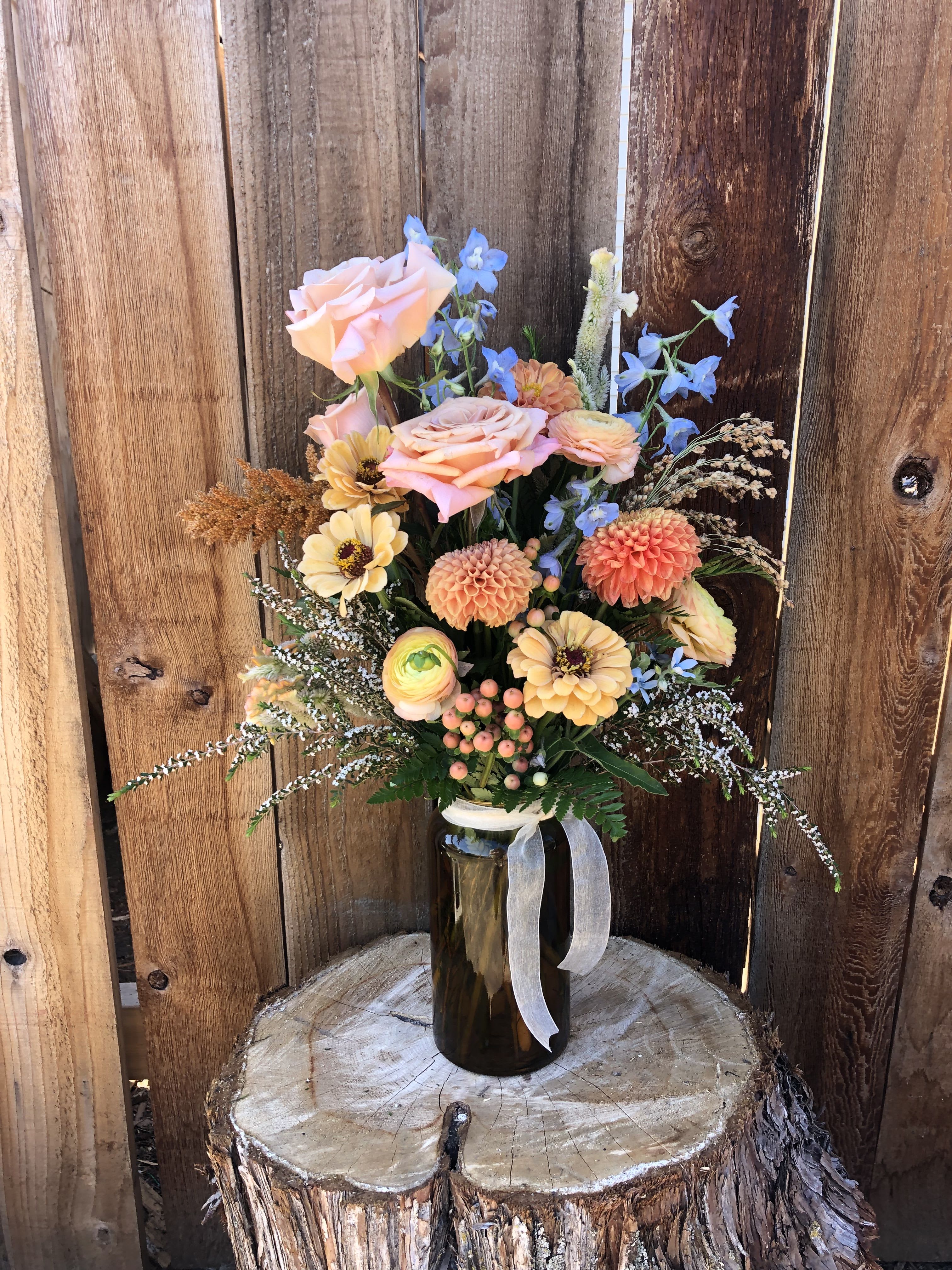 Rustic arrangement of peach roses, dahlias and mixed blooms in an amber glass jar with ribbon.