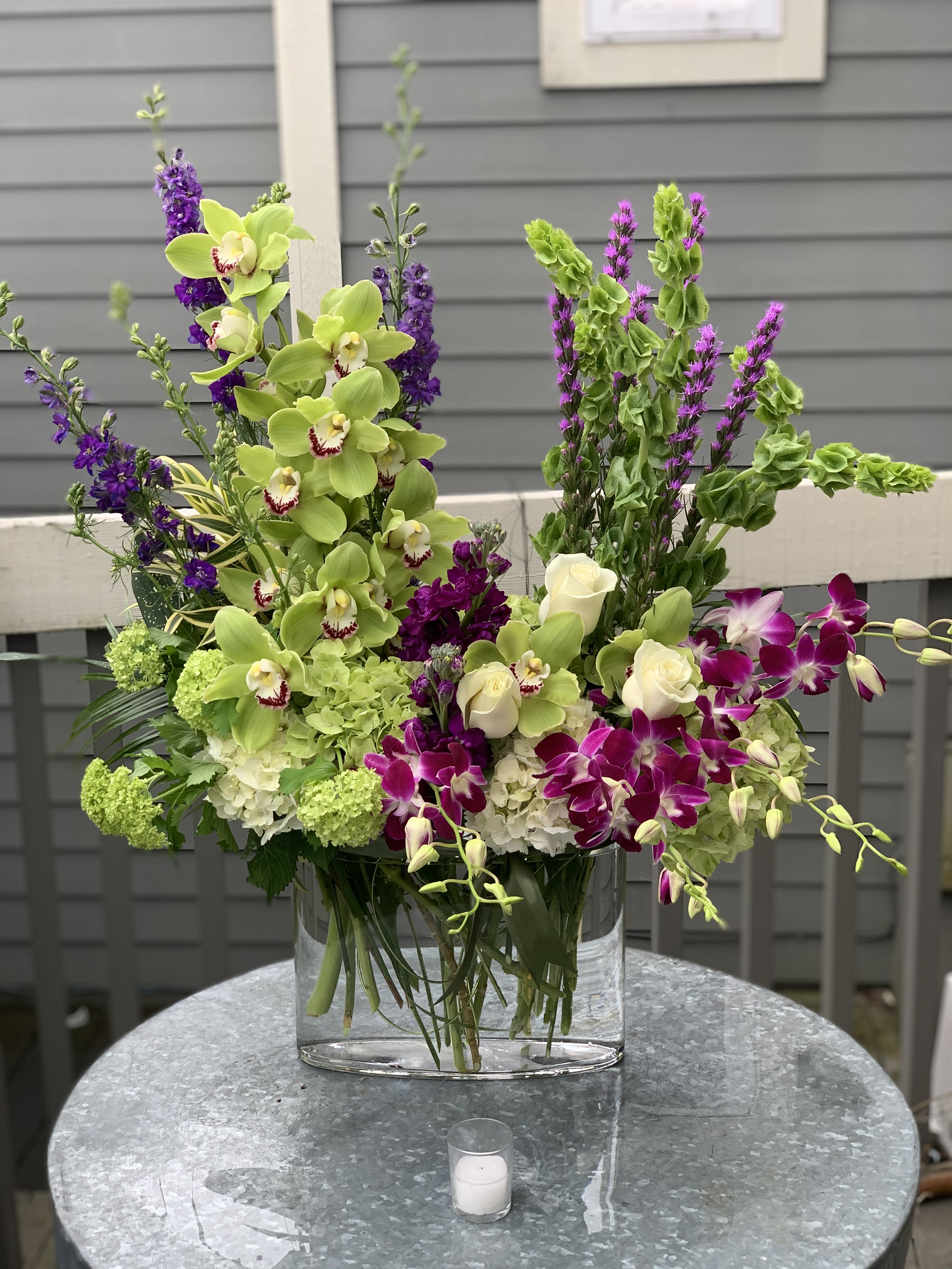 Tall floral arrangement with green orchids, purple blooms, and white roses in a glass vase