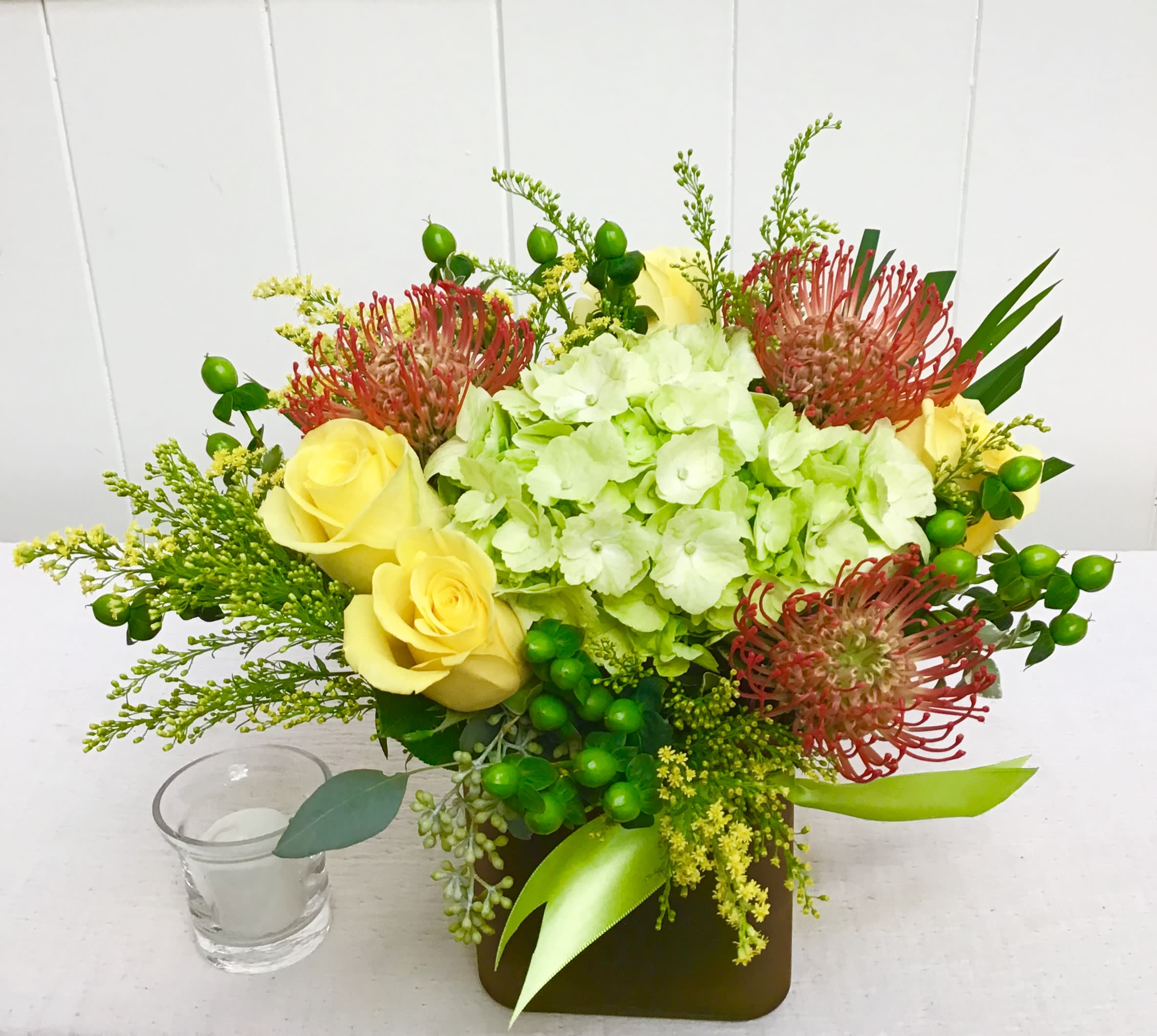 Low arrangement of green hydrangea, yellow roses, and red pincushion protea in a square vase with a small votive candle.