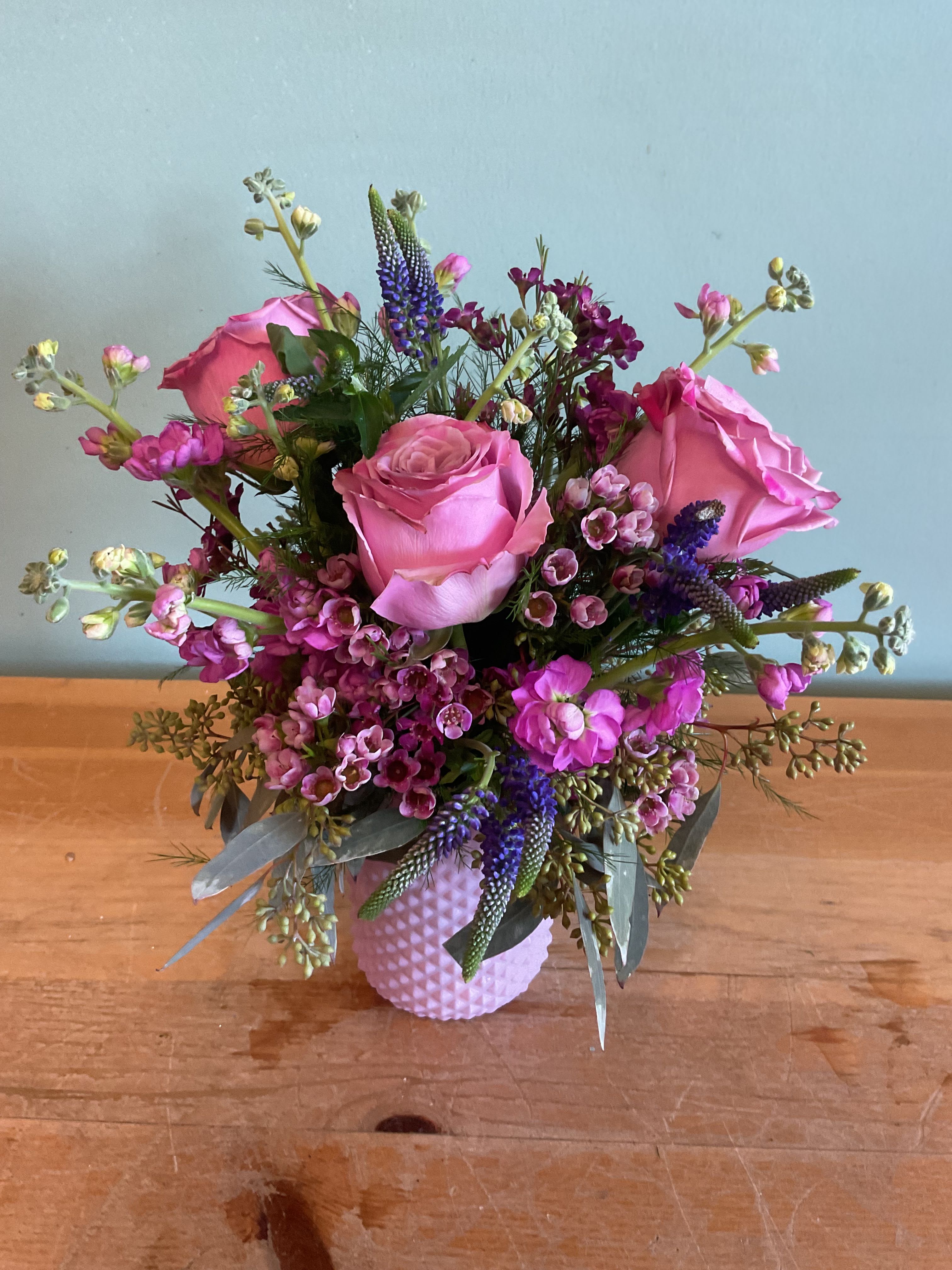Neat &amp; Petite - A cluster of soft pink roses takes center stage in this Neat &amp; Petite flower arrangement, making it an ideal birthday bouquet, thank-you gift, or everyday surprise for someone who loves feminine, romantic color. Upright purple spikes and sprigs of magenta and lavender filler flowers give the arrangement a lively, garden-style silhouette that feels cheerful and slightly whimsical-perfect for marking a small celebration or letting someone know you're thinking of them.  The design is compact but full, arranged in a textured pink ceramic vase that complements the pink, purple, white, and green palette. Expect rounded, outward-reaching stems that create movement and texture alongside classic rose blooms and fragrant stock. Sized to sit comfortably on a bedside table or desk, this petite arrangement brightens small spaces and arrives ready to display with minimal care-fresh water and a clean vase keep these blooms looking their best.