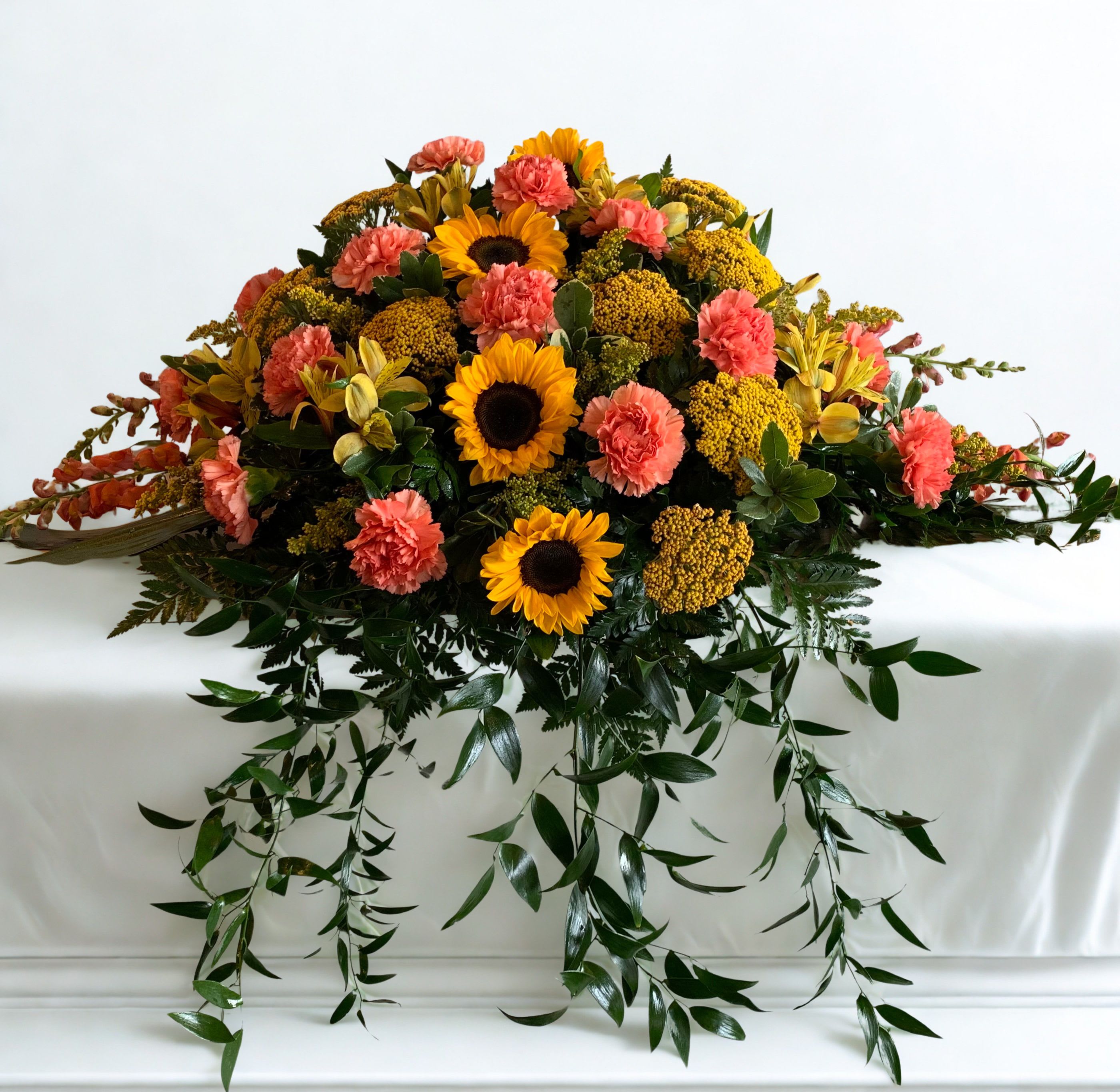 Eternal Sunshine - Bright yellow sunflowers paired with coral-pink carnations make a warm, uplifting sympathy spray that brings sunny color to a memorial or service. The low, wide shape reads as a formal tribute while still feeling hopeful-perfect for honoring a life with cheerful, consoling blooms that say you're thinking of them.  This large, low floral spray features clustered sunflowers and coral-pink carnations across the top, accented by rounded yellow filler blooms for texture and fullness. Long, trailing greenery spills downward for a gentle cascade, giving the arrangement presence on a table or casket display. The palette of yellow, pink and fresh green creates an abundant, comforting impression without being overstated.  Designed for sympathy or memorial display, this arrangement offers a heartfelt way to celebrate a life with warmth and light. If you need it sized or delivered specifically for a service, please select the appropriate delivery option at checkout or contact us for timing to ensure it arrives for the ceremony.  As similar as possible. Designer may make appropriate changes based on flower availability. 