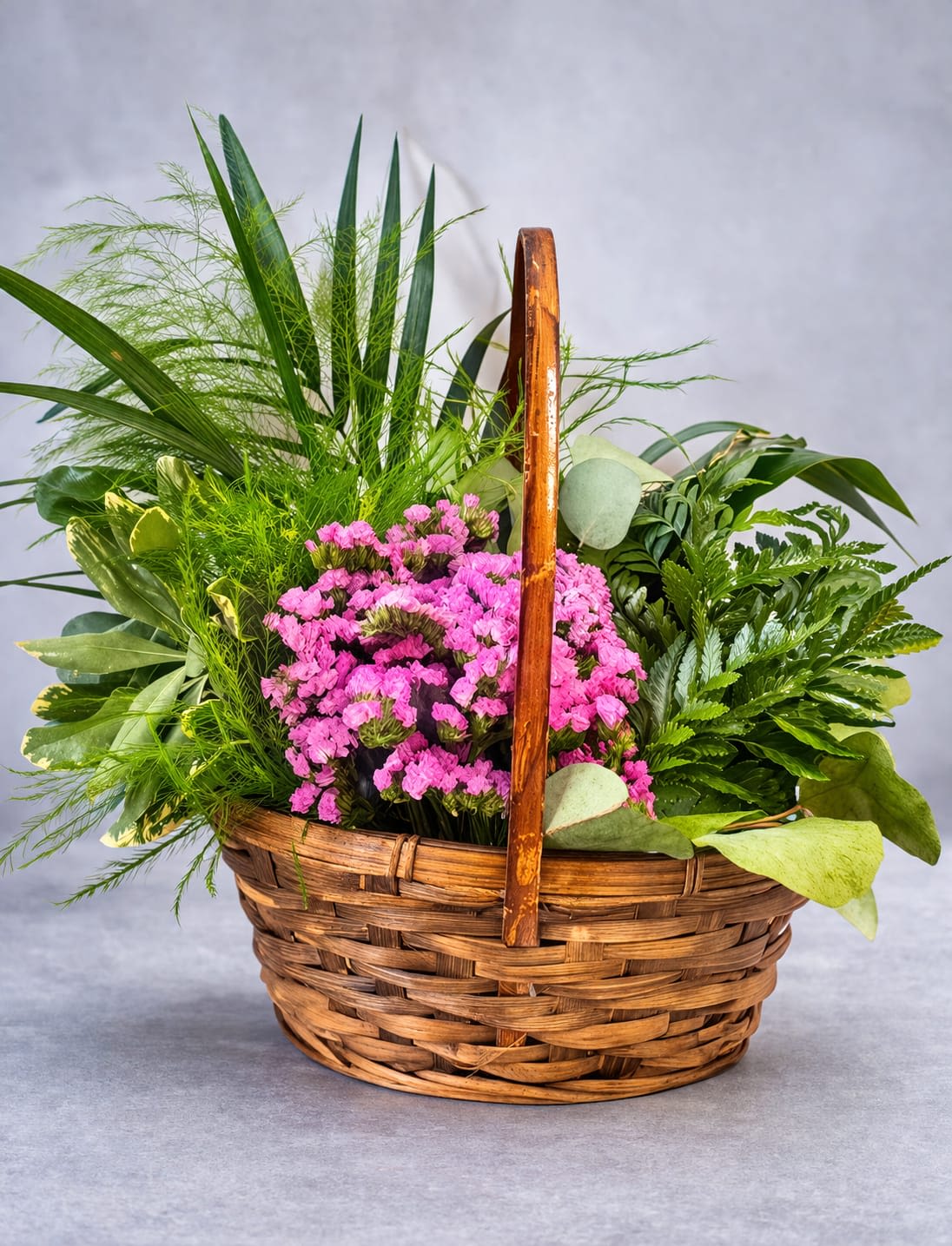 Garden Harmony - Bright pink carnations burst from a low wicker basket in this Garden Harmony flower arrangement, making it an ideal birthday bouquet, thank-you gift, or "thinking of you" surprise. The vivid pink blooms form a dense, cheerful cluster so the person you're celebrating will feel noticed and genuinely uplifted the moment it arrives.  Surrounded by varied greenery-long feathery fronds and broader leaves-the flowers sit in a warm brown wicker basket with a handle that gives the design a natural, rustic cottage-garden look. Low and full, the arrangement reads as an everyday classic that fits on a table or desk; arrange in a cool spot and change the water regularly to keep carnations fresh longer.
