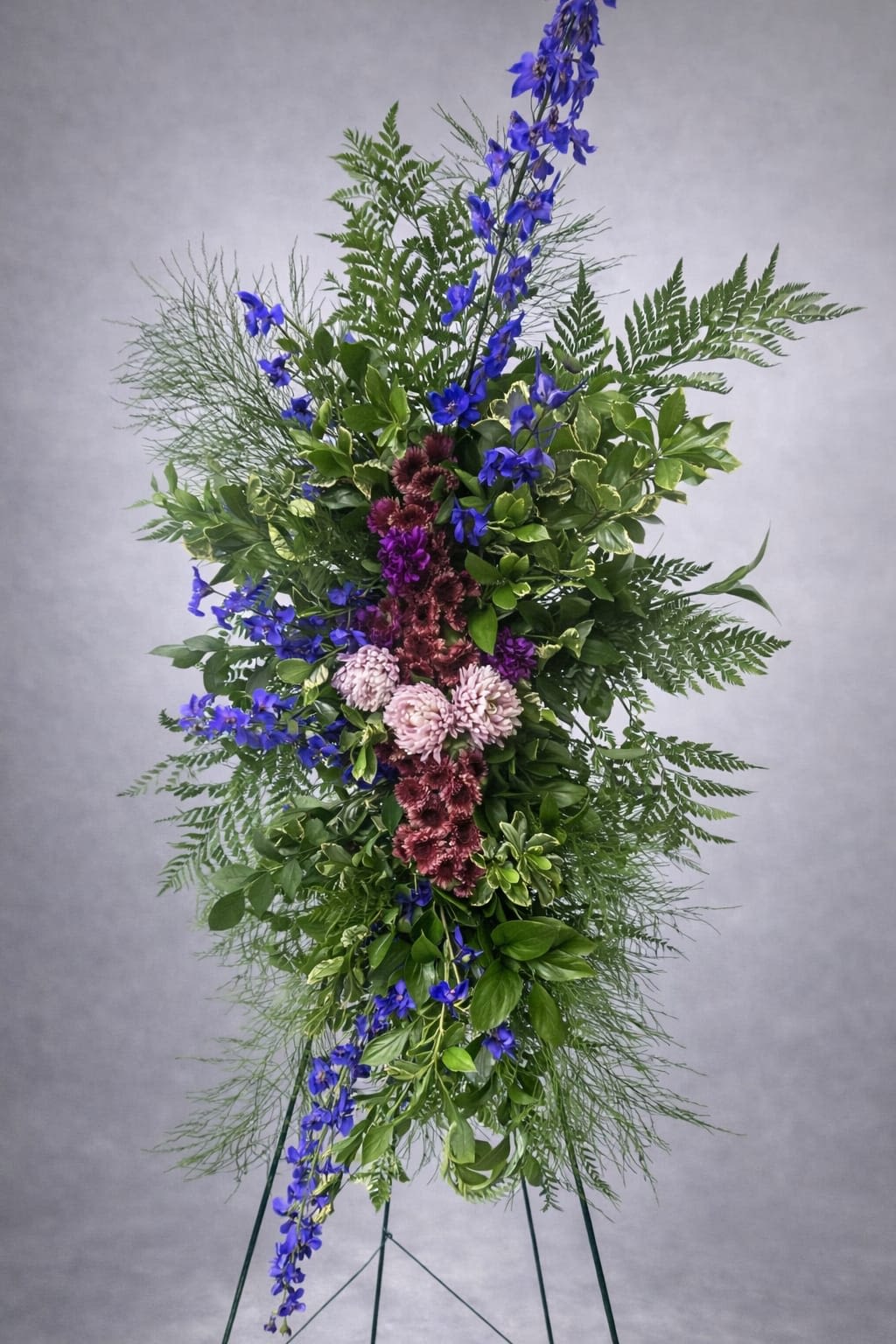 Eternal Peace Standing Spray - A tall, cool-toned standing spray that reads as a dignified tribute - perfect for sympathy or a memorial service. The vertical composition centers vivid blue delphinium-like spikes that lift the eye, while clusters of purple and blush-pink chrysanthemums bring soft color and textured form. This arrangement communicates calm and remembrance, the kind of expression that helps the person you're honoring feel seen.  Dense mixed greenery and airy fern fronds create a layered, open silhouette that keeps the spray from feeling heavy; fine foliage weaves between blooms for a natural, flowing look. Sized for formal presentation, it arrives mounted on a metal easel for immediate display at services, funeral homes, or graveside ceremonies. Care is minimal - keep the floral foam watered if displayed for longer periods to maintain freshness.