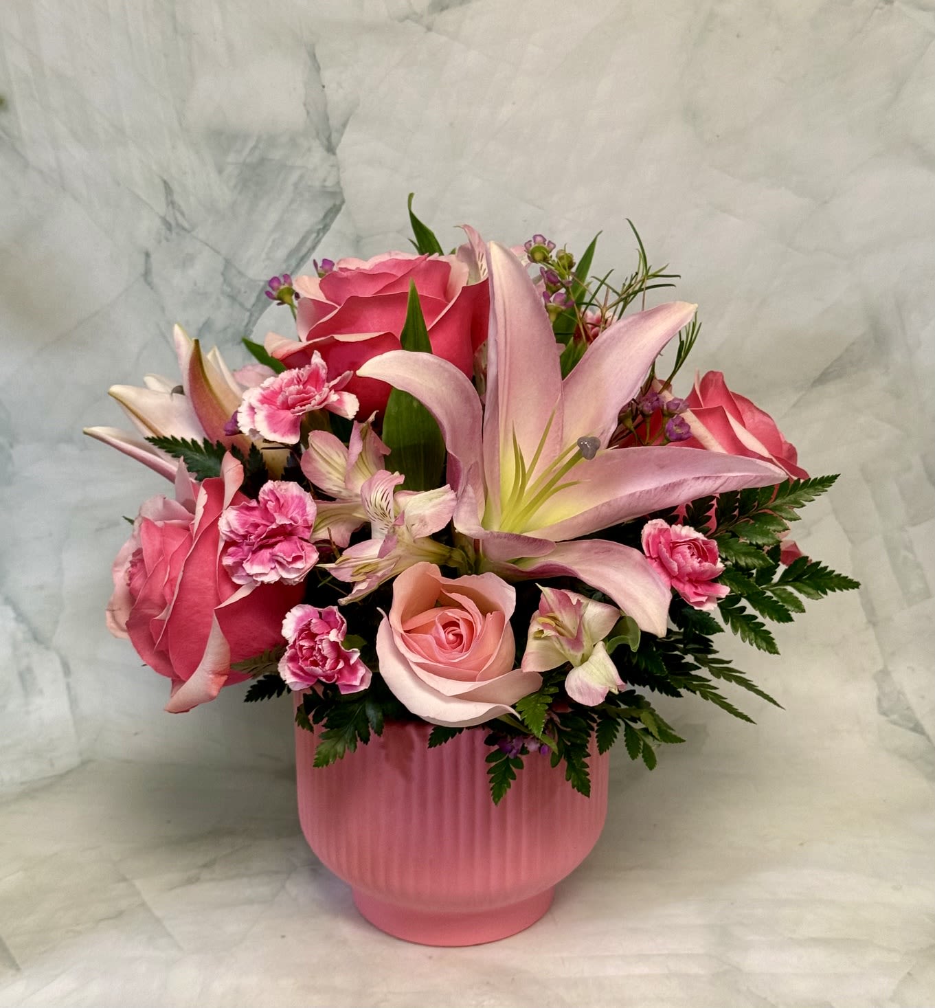 The Modern Mom - Lush pink lilies and soft blush roses take center stage in this spring-ready flower arrangement designed for Mother's Day, birthdays, or "just because" moments. The Modern Mom arrangement reads romantic and cheerful-the big, scented lilies and layered roses are softened by ruffled pink carnations and small purple filler blooms so the person you're thinking of will feel truly celebrated when it arrives.  Arranged in a ribbed pink vase, the bouquet blends pink, white, green, and touches of purple for a full, rounded silhouette that suits a mantel, dining table, or bedside. Hydrangeas, chrysanthemums, alstroemeria, and fresh greenery add texture and volume around the focal lilies and roses, creating a garden-inspired mix with lively magenta accents. Handcrafted in Philadelphia, it's an elegant everyday gift that looks abundant without feeling fussy.  This piece is sized to be a standout centerpiece yet easy to display-perfect for springtime gatherings or sending thanks to a mom who deserves something special. For best enjoyment, trim stems and refresh the water every couple of days to keep blooms looking their freshest.