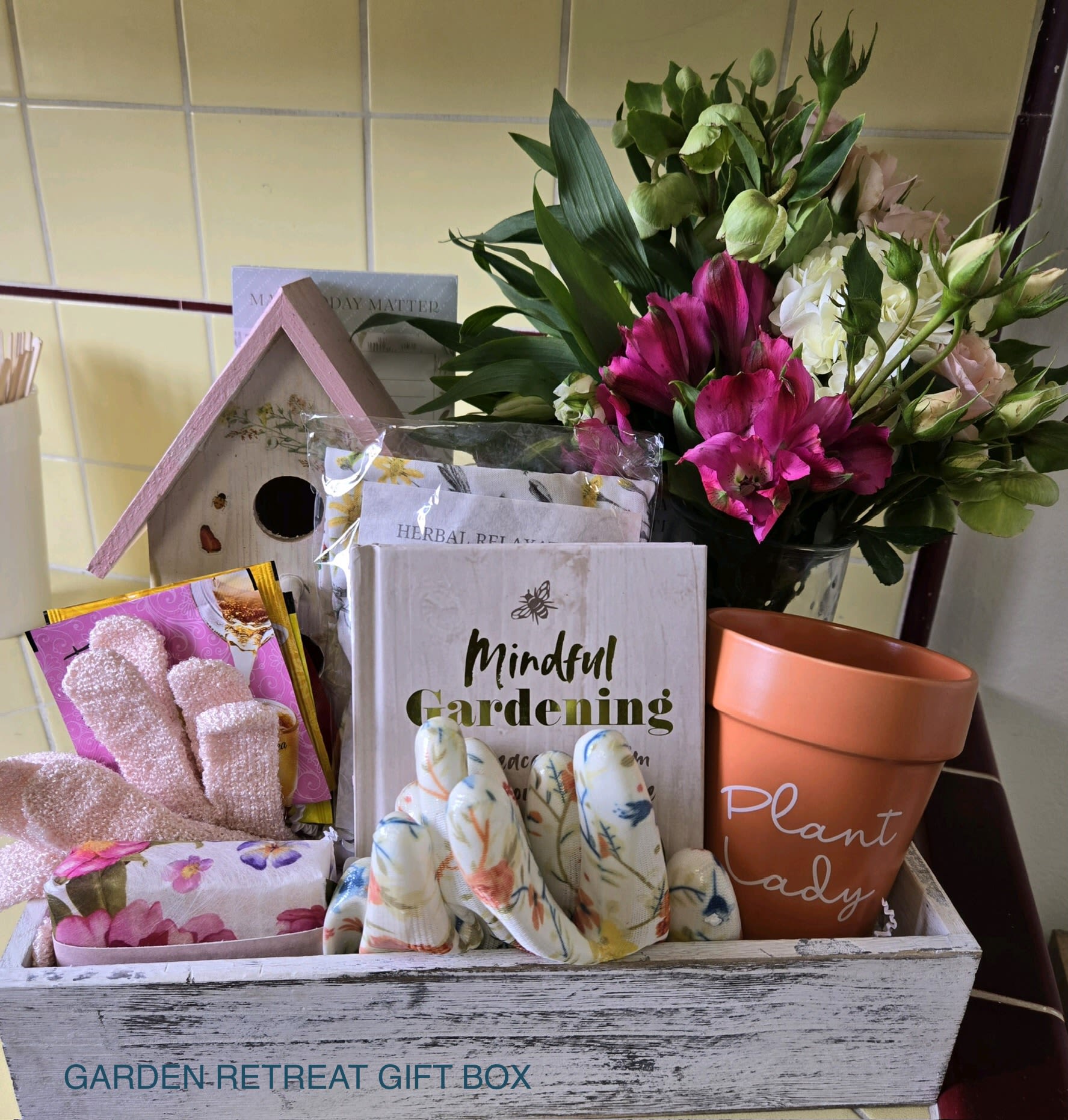 Garden Retreat Gift Box - A lively mixed bouquet paired with thoughtful garden-themed gifts makes this wooden gift box an ideal present for a plant-lover or someone tending a new garden. This gift set reads as a garden celebration and is perfect for birthdays, Mother's Day, or a "thinking of you" surprise-the bright pink alstroemeria and soft white lisianthus headline a feminine palette of magenta, blush and green that feels both joyful and personal.  Arranged in a rustic wooden crate, the bouquet's dark green foliage frames vivid magenta alstroemeria, pale white lisianthus and complementary blooms for texture and movement. Alongside the flowers you'll find curated hard goods that reinforce the hobby-first theme: a terracotta plant pot labeled "Plant Lady," patterned garden gloves, a small decorative birdhouse, a gardening book, and wrapped seed packets-everything presented for an unboxing that delights the recipient.  Give this to the person who loves potting, porch gardens, or simply the ritual of caring for plants; it says "I see your passion" more clearly than a vase alone. The crate is ready to display or repurpose for storage, and the included items make it an immediately useful and heartwarming gift-no extra shopping required.