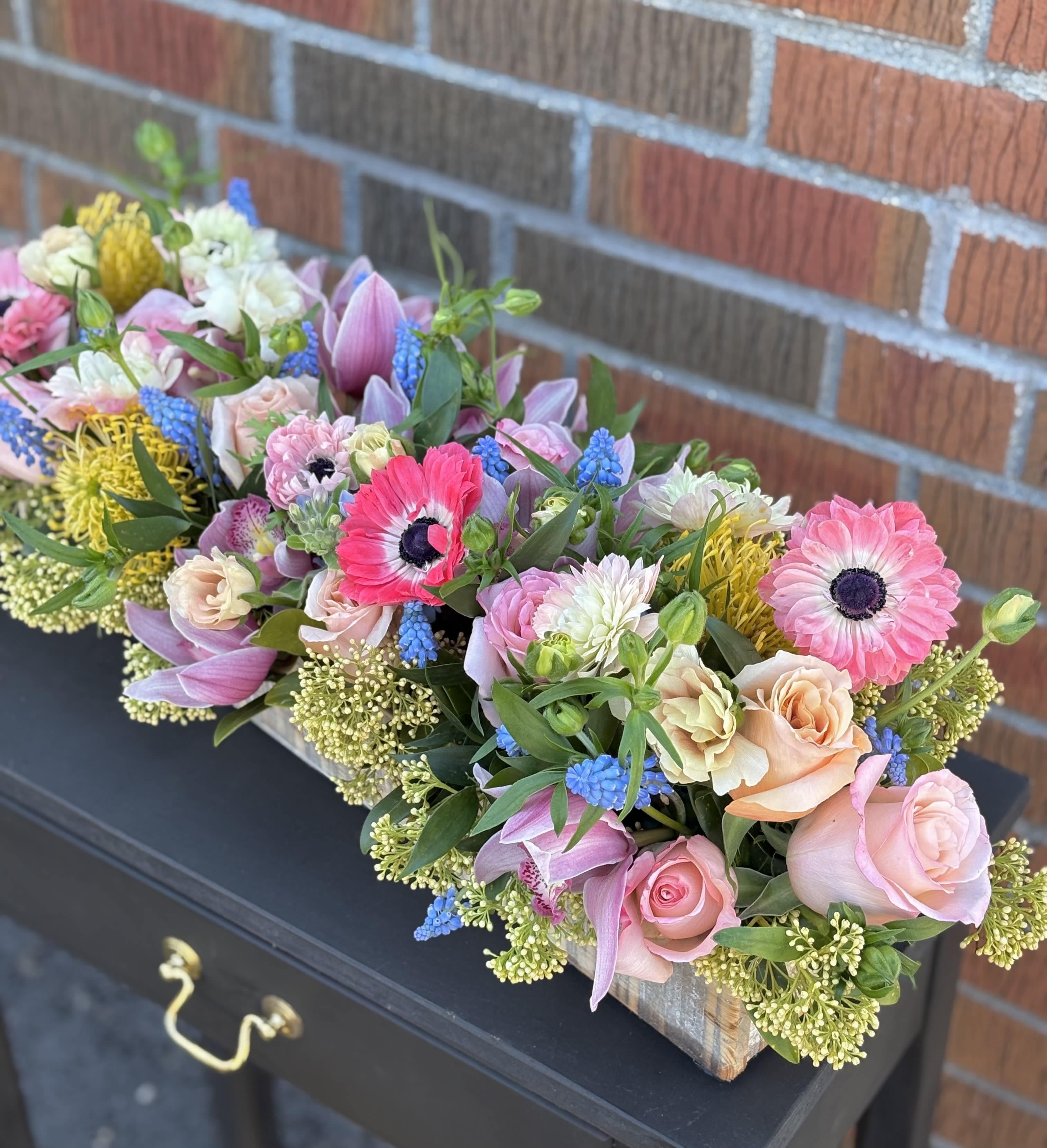 Long wooden box arrangement with pink gerbera daisies, roses, dahlias, blue hyacinths, and yellow protea.