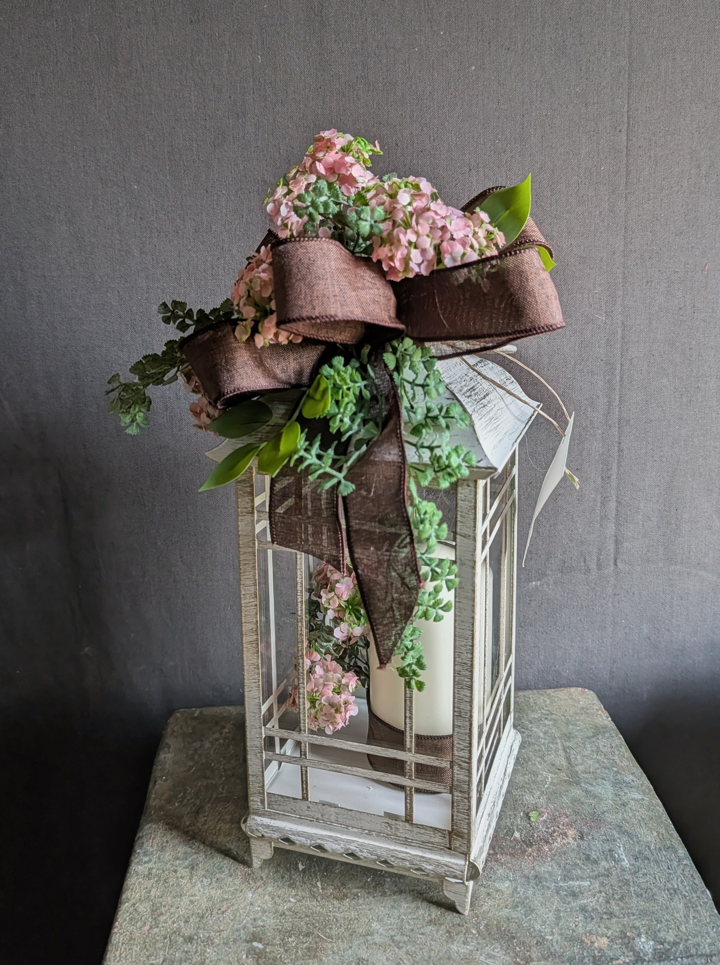 Garden Lantern Centerpiece - Soft pink blooms nestled around a tall decorative lantern create a romantic centerpiece that reads like a thoughtful gift-perfect as a birthday bouquet alternative, a housewarming surprise, or a "thinking of you" token. The lantern and decorative candle inside give a clean, gentle show that contrasts with the fuller floral top and trailing accents, so the person you're sending it to will feel warmly celebrated and comforted whether it's for everyday display or a quieter sympathy moment.  This maintenance-free arrangement combines mixed faux blooms and greenery gathered tightly atop and around the lantern. A wide brown ribbon is tied in layered loops with a long tail for a rustic, gift-ready finish; the cream candle sits inside the metal lantern for a soft visual candlelight effect. Sized to dress entry tables, mantels, and dining room centers, it's an easy, lasting way to mark birthdays, housewarmings, Earth Day displays, gift sets, or to offer gentle condolences-no watering or replacement needed.
