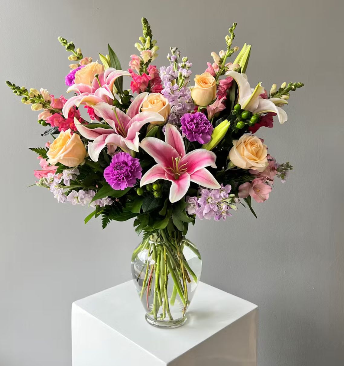 Blushing Elegance Bouquet - The Blushing Elegance Bouquet is a radiant blend of charm and sophistication. Featuring fragrant pink lilies, soft peach roses, and vivid purple blooms, this arrangement captures the beauty of spring in every petal. Artfully arranged in a clear glass vase, it’s perfect for celebrating birthdays, anniversaries, or simply brightening someone’s day with a touch of floral grace.