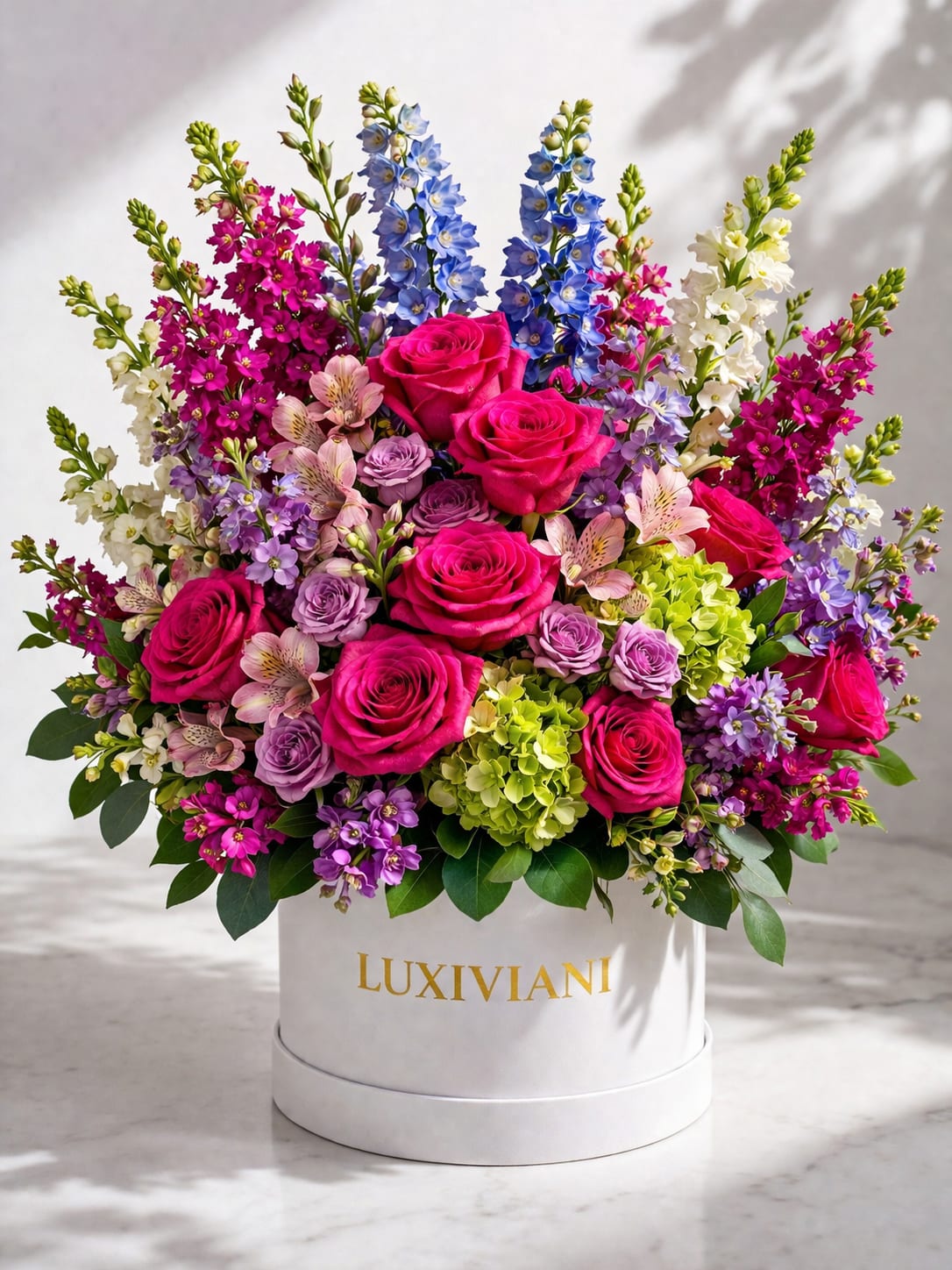 The Signature Bloom - Vivid pink and lavender roses take center stage in this romantic birthday bouquet, rising from a polished white hat box for an upscale presentation. The Signature Bloom reads as a celebration in color-bright pinks and purples softened by blue and white spires-so the person you're thinking of will feel noticed and adored the moment it arrives.  This compact, rounded arrangement mixes classic roses with alstroemeria, fragrant stock, and a lime-green hydrangea cluster for lively contrast and layered texture. Tall blue and white floral spires add vertical movement above the densely packed blooms, all arranged in a white cylindrical hat box with gold lettering for easy display and gifting.  Perfect for anniversaries, birthdays, or simply saying "I love you," this arrangement arrives ready to set on a table or counter-no vase required. For best enjoyment, keep the box in a cool spot out of direct sun and refresh any visible water source if prompted by seasonal care instructions.