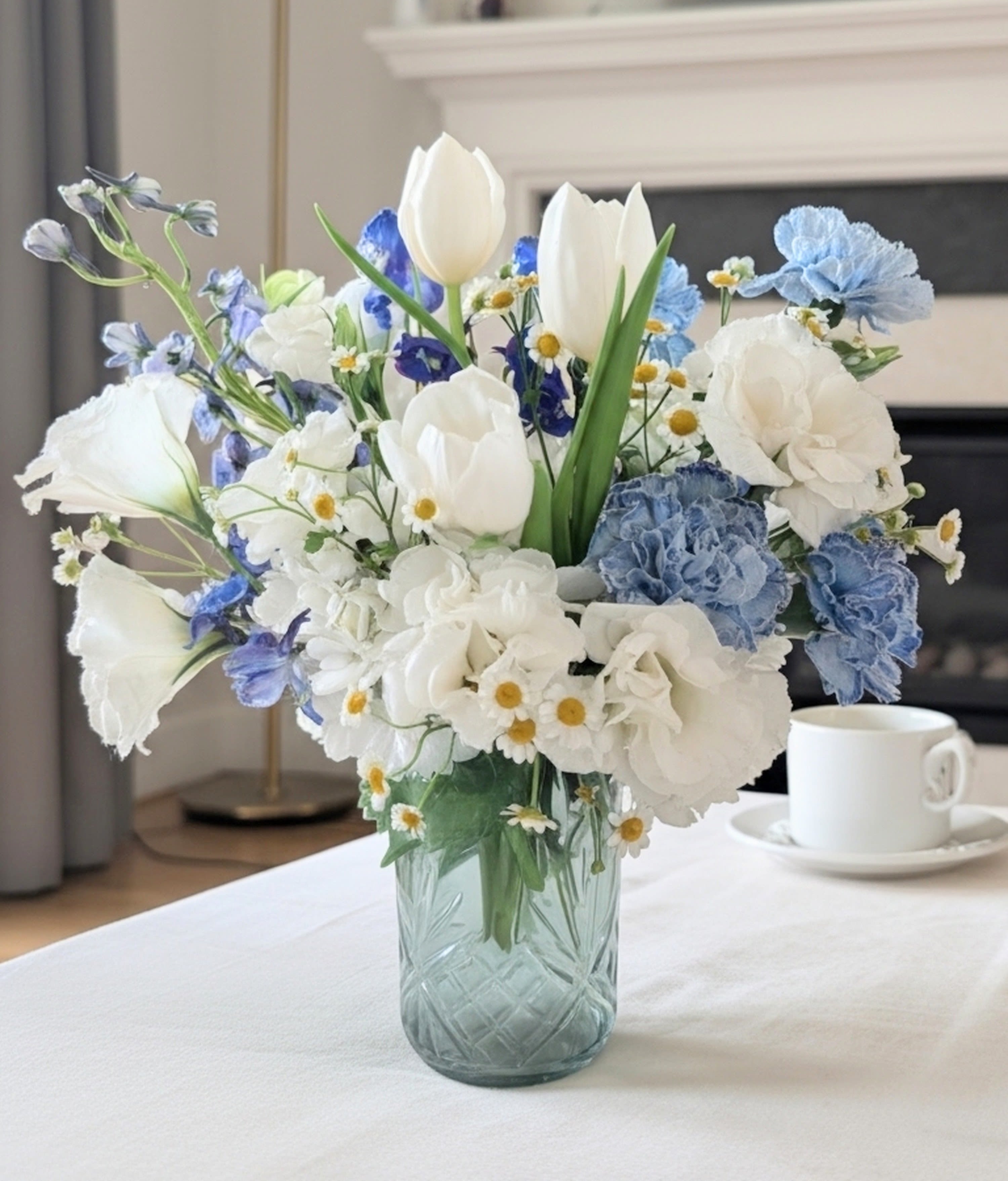 Serene Skies Bouquet - White tulips and lisianthus plus small clusters of chamomile-like blooms mingle with soft blue carnations and cool blue accents to create a calm, airy flower arrangement perfect for a spring gift or a housewarming bouquet. As a flower arrangement for everyday gifting or to mark a quiet celebration, Serene Skies Bouquet sends a message of peaceful, thoughtful care - the kind of display that makes the person you're thinking of feel gently seen and comforted.  The composition is full and rounded: taller tulip stems rise above a cloud of softer blooms that spread outward, giving the bouquet a natural, garden-fresh silhouette. Hues of white, blue, and green combine for an elegant, restrained palette; a clear cut-glass vase anchors the design and adds classic home-decor charm. 