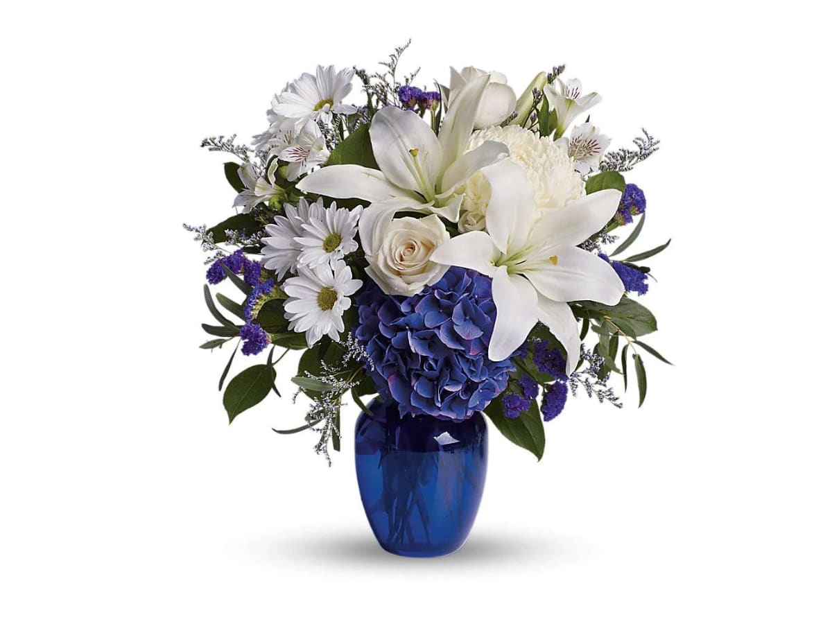 BEAUTIFUL IN BLUE - In this arrangement, the serenity of the color blue along with the purity of intention symbolized by white will let the family know you are sending your calm strength to them during these difficult times.  As shown: Designed in a 9" vase.  Deluxe and Premium made in an 11" vase.  Beautiful blooms such as blue hydrangea, crème roses, white lilies and alstroemeria along with yellow and white chrysanthemums, eucalyptus, limonium and more are beautifully arranged in a dazzling cobalt blue vase. Approximately 16 1/2" W x 18" H