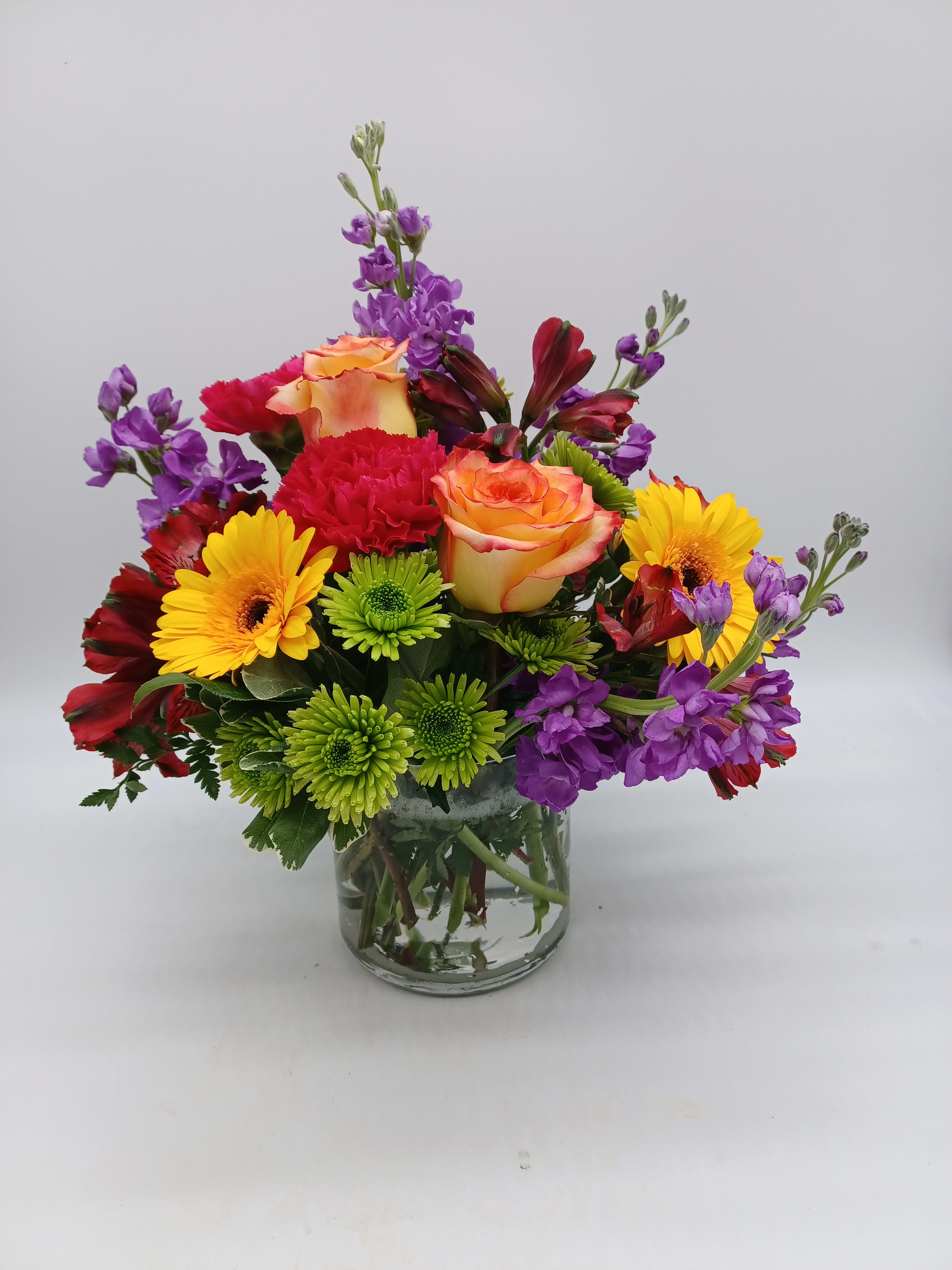 Mixed bouquet of roses, gerbera daisies, carnations, and purple blooms in a glass vase