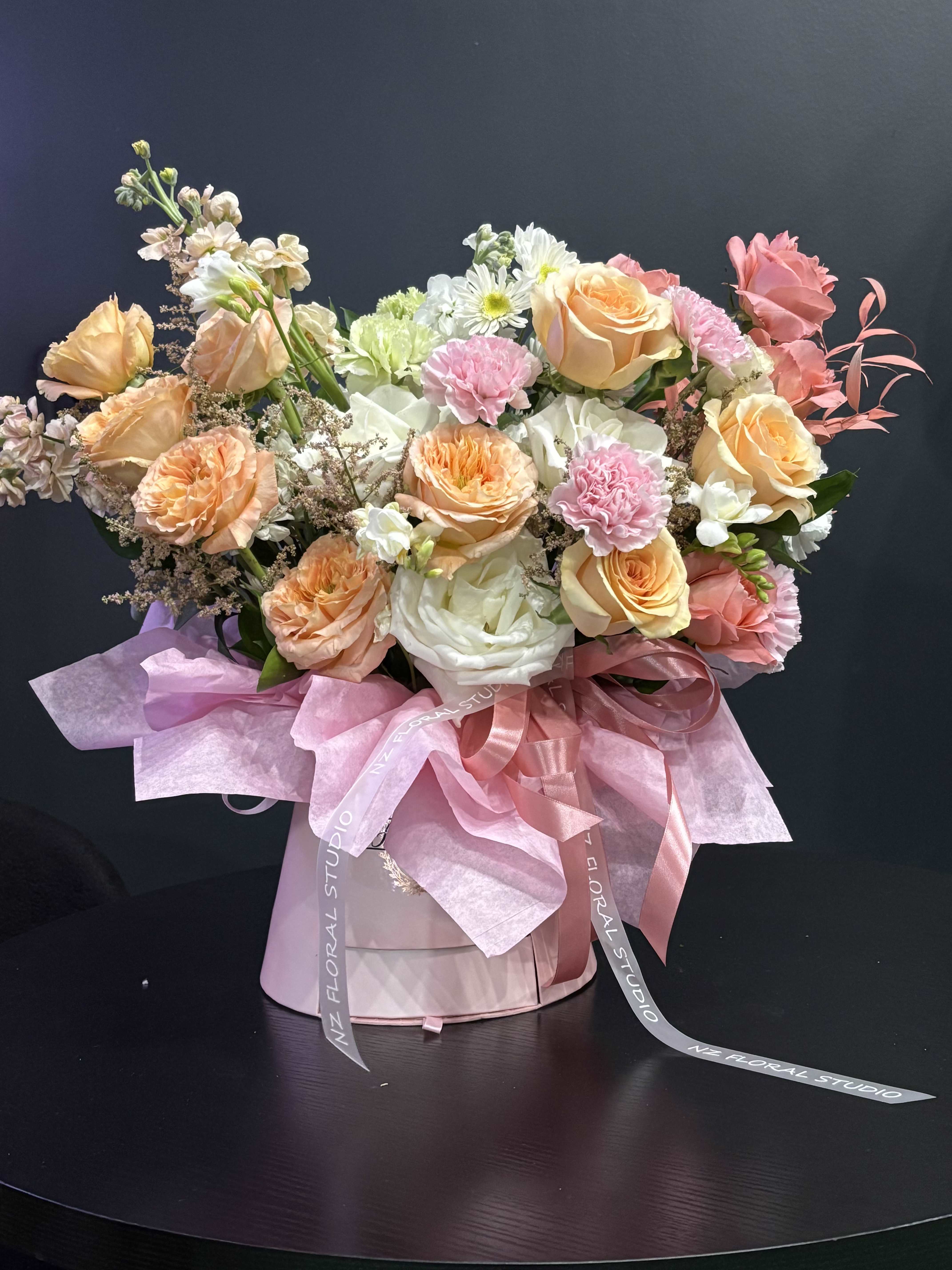 Blush Garden - Blush Garden is a delicate, garden-inspired arrangement designed in soft pastel tones to create a feeling of lightness, beauty, and quiet elegance. Featuring a harmonious blend of blush and peach roses, airy chamomile, and refined white and lavender chrysanthemums, this composition feels both fresh and naturally sophisticated. Each bloom is thoughtfully placed to create gentle movement and an organic shape, giving the arrangement a soft, effortless look — as if gathered from a blooming garden. The combination of textures and subtle color transitions adds depth while maintaining a calm, romantic aesthetic. Presented in an elegant hat box with flowing ribbon details, Blush Garden is perfect for meaningful moments, heartfelt gestures, or simply bringing a touch of beauty into everyday life. This design is created for those who appreciate softness, femininity, and understated luxury.