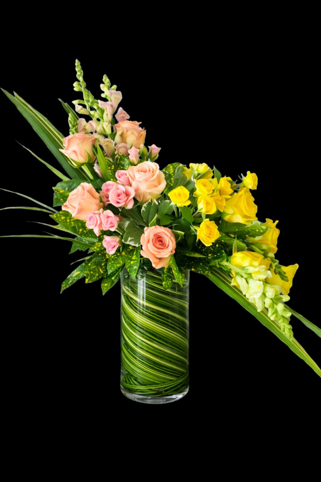 Spring Awakening - Peach, pink and sunny yellow roses spill from a tall clear glass vase in a bright, cheerful spring bouquet-perfect as a birthday bouquet, a thank-you arrangement, or a pick-me-up for someone celebrating the season. A few pale snapdragon spikes rise above the roses to add graceful height and movement, so the person you send this to will feel seen and refreshed by its lively, uplifting presence.  Tightly layered green foliage and long leaves fill the vase, creating a fresh, verdant base that makes the warm rose tones pop. The design is full and slightly asymmetrical, with concentrated yellow roses on one side balanced by softer peach and pink blooms at center and left for natural charm. The clear glass vase showcases the stems and layered greenery for a clean, modern look that fits mantelpieces, desks, or kitchen tables.  This arrangement suits spring celebrations, everyday gifting, or sending thanks-any moment when you want the recipient to feel genuinely celebrated. Vase dimensions and care: keep stems in fresh water, recut stems every few days, and place away from direct heat to prolong bloom life.