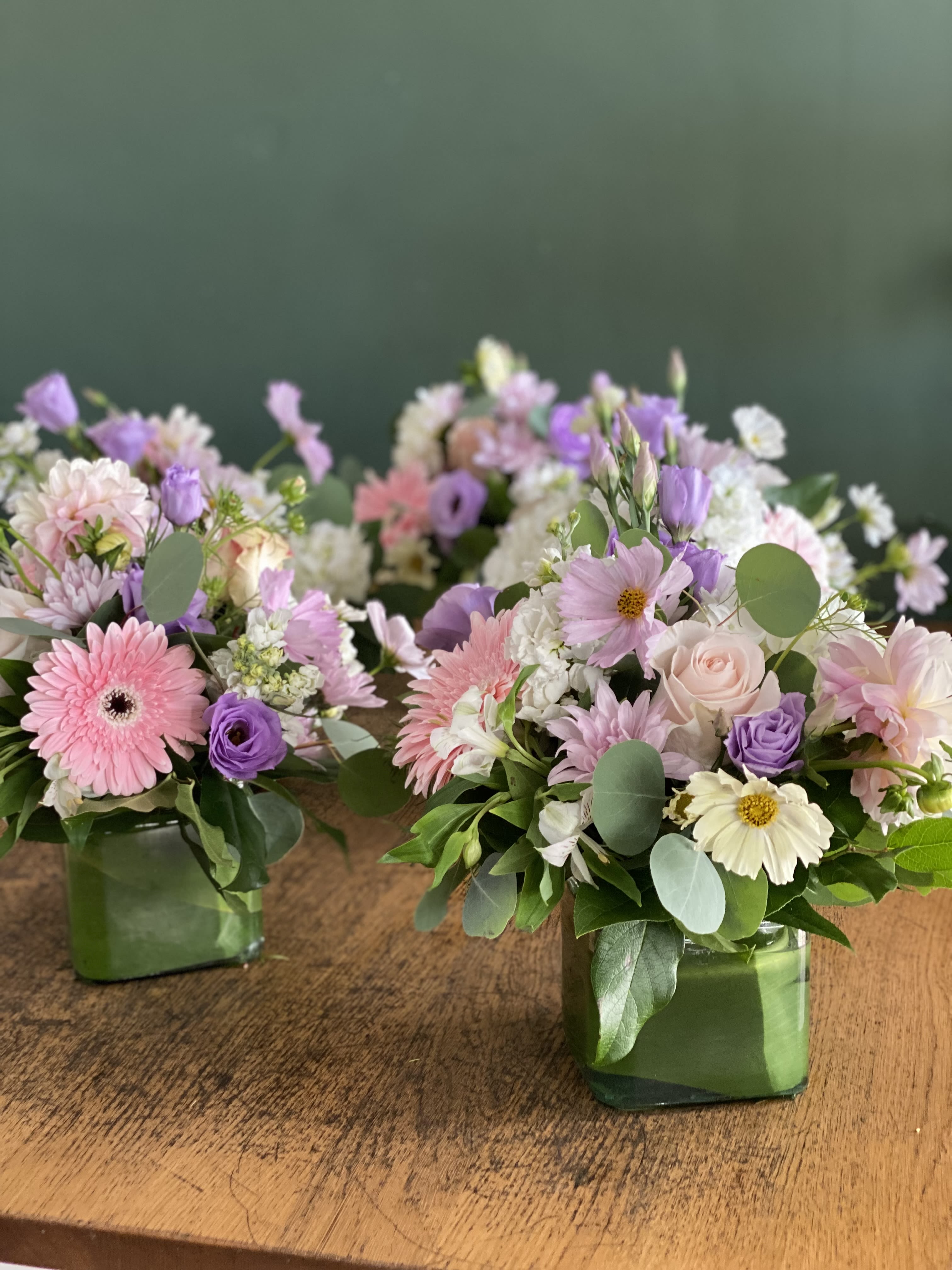Two pastel floral arrangements in square glass vases on a wooden table