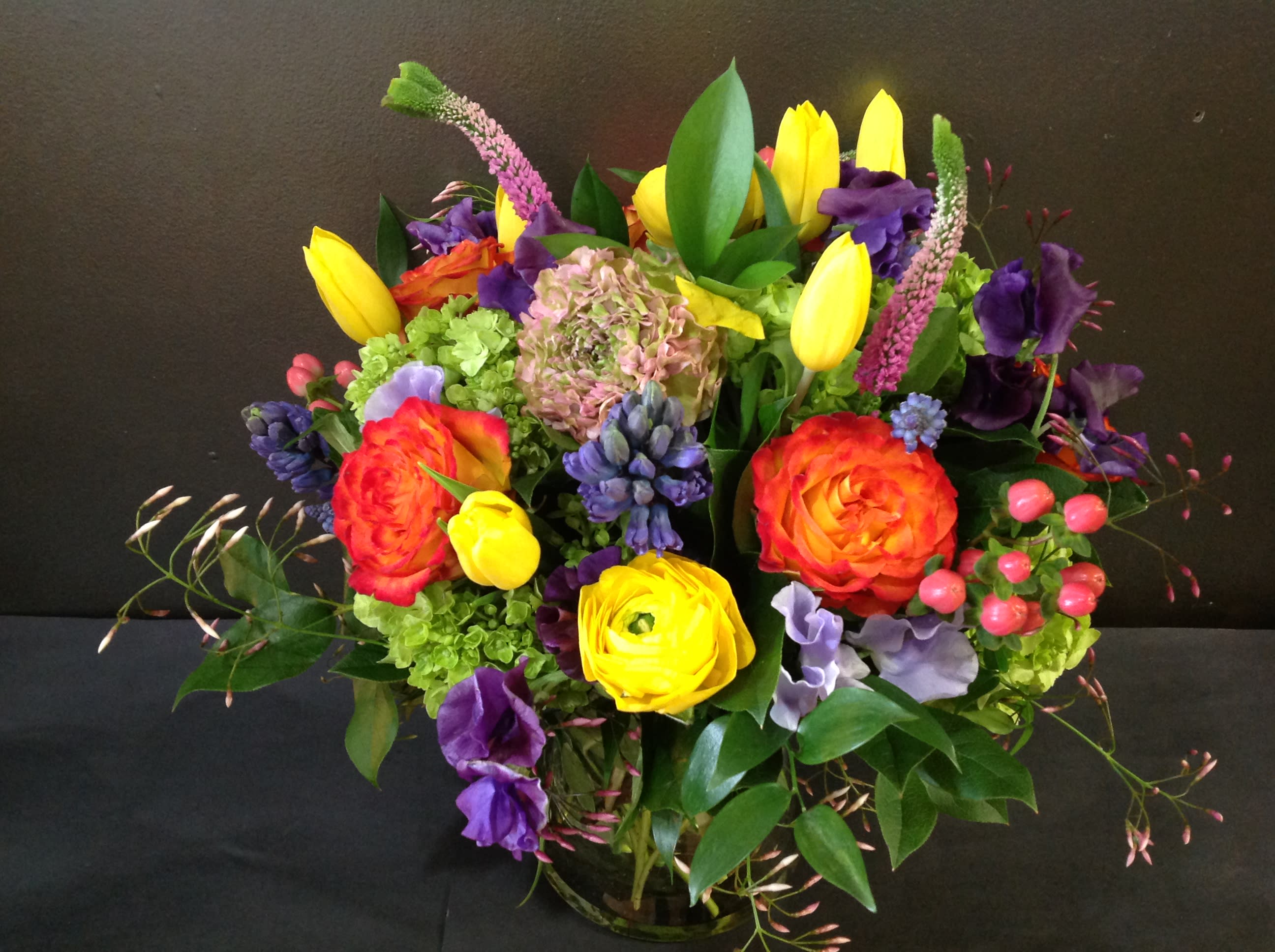 Bright mixed arrangement of yellow tulips, orange roses and purple blooms in a clear glass vase