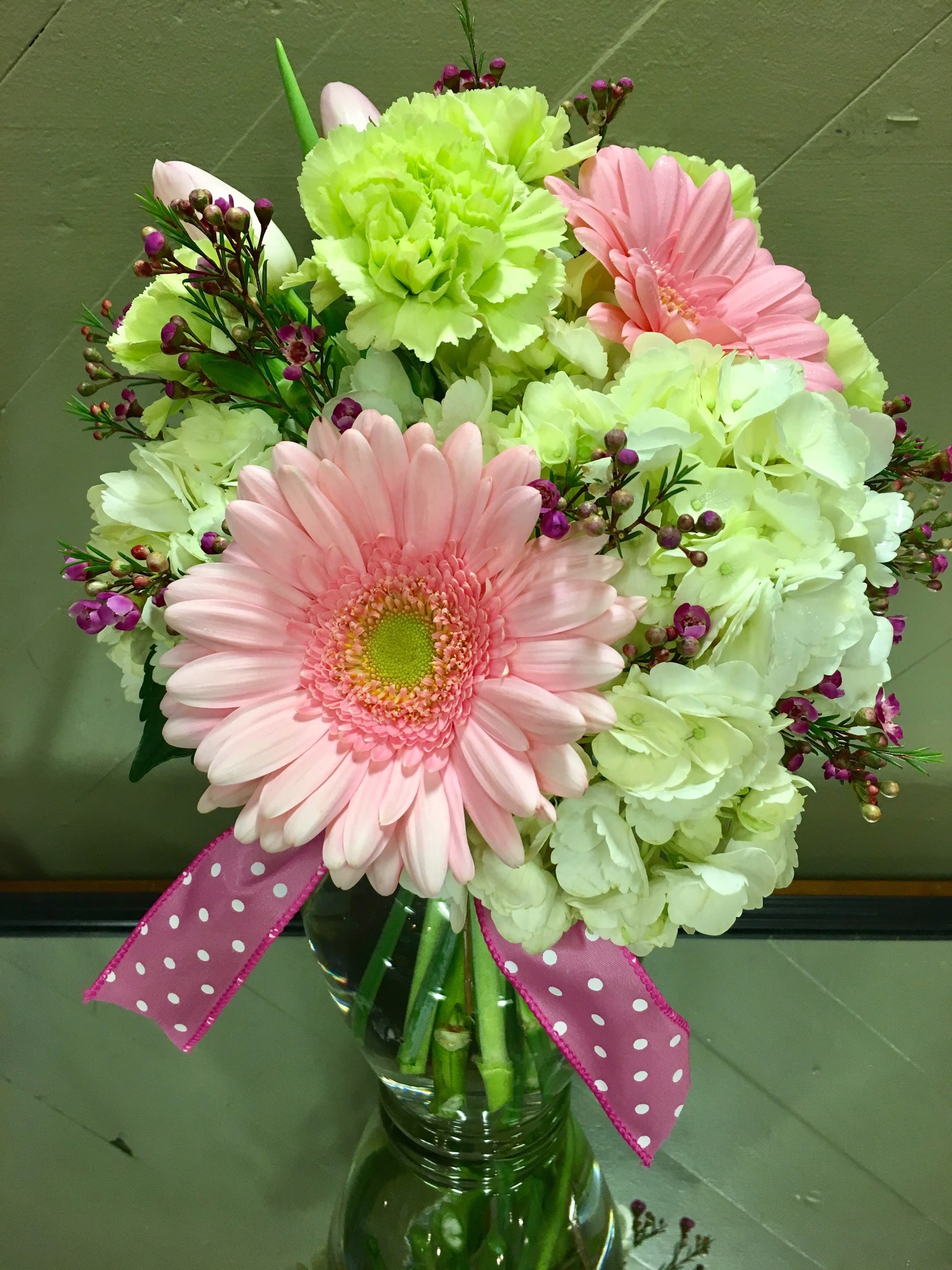 Pink gerbera daisies and pale green carnations in a glass vase with a polka-dot ribbon.
